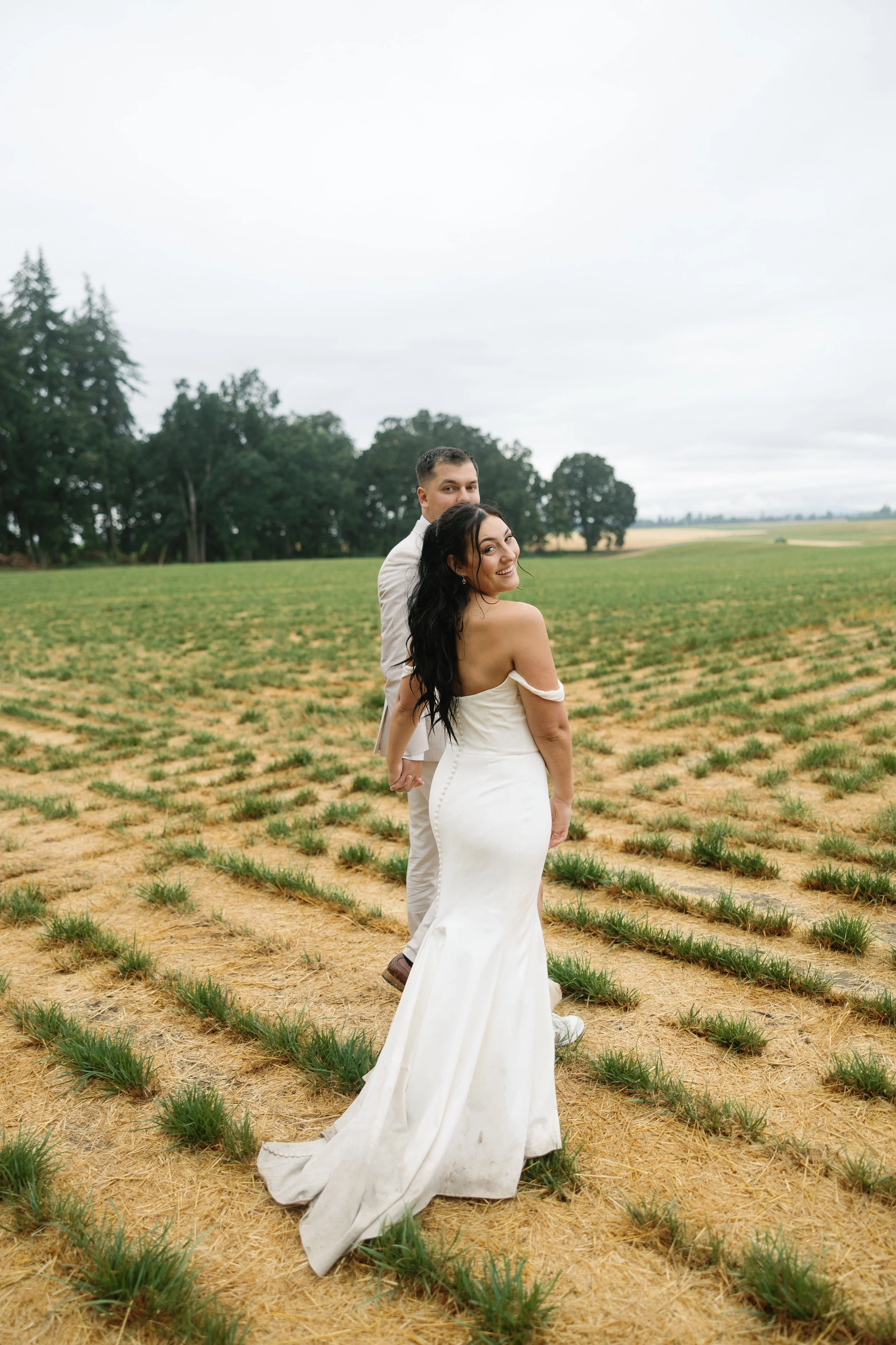 A bride and groom holding hands in an open field, with the bride in a white wedding gown and the groom in a light-colored suit, smiling at the camera on a cloudy day.