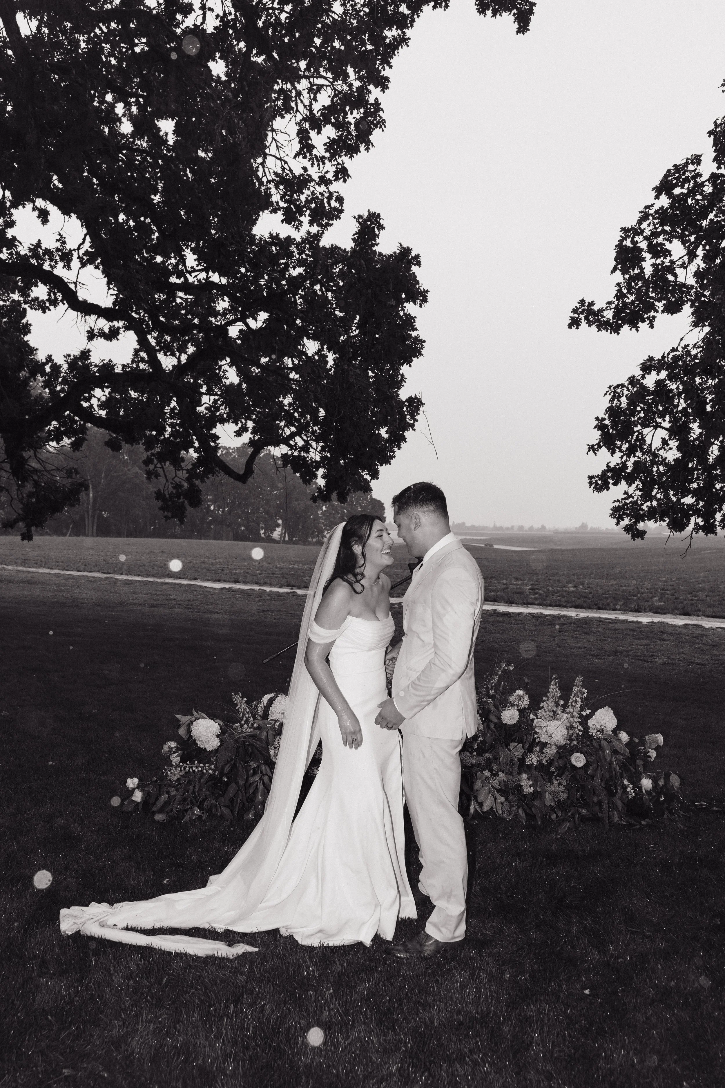 A couple in wedding attire standing and smiling close together outdoors, with trees and an open field in the background, under a large tree, in black and white.