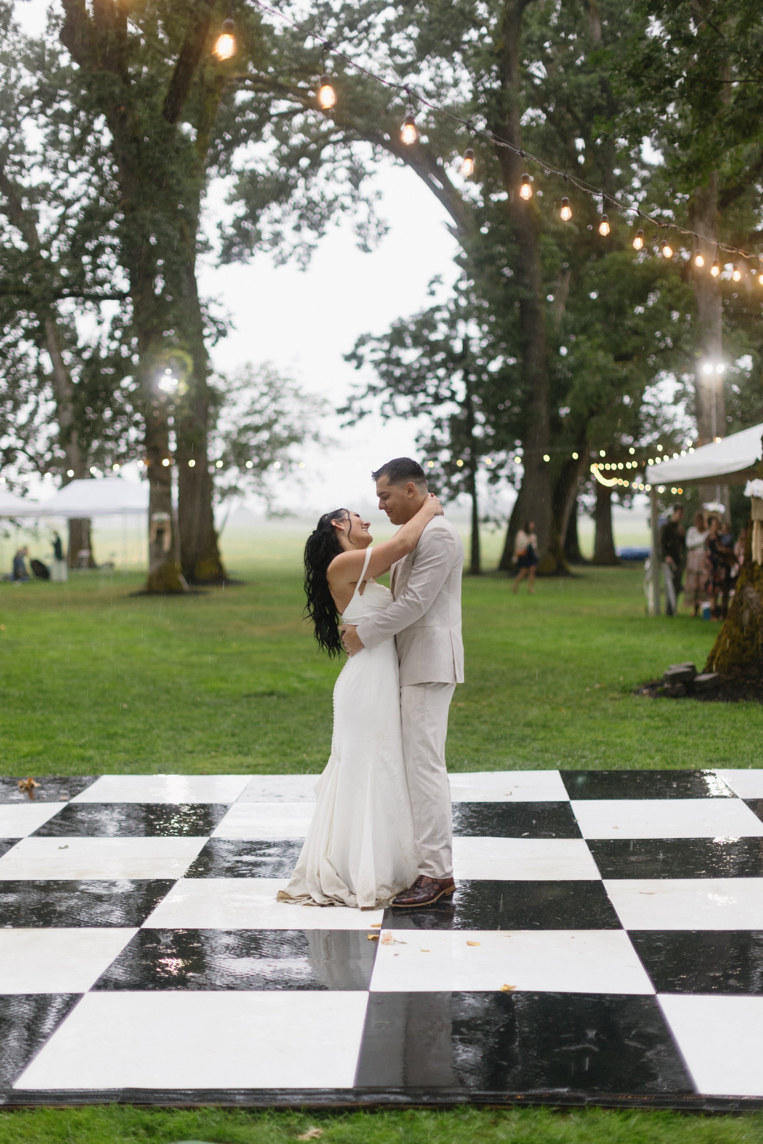 Couple dancing on a black and white checkered dance floor outdoors, surrounded by trees and string lights, during an evening wedding reception.