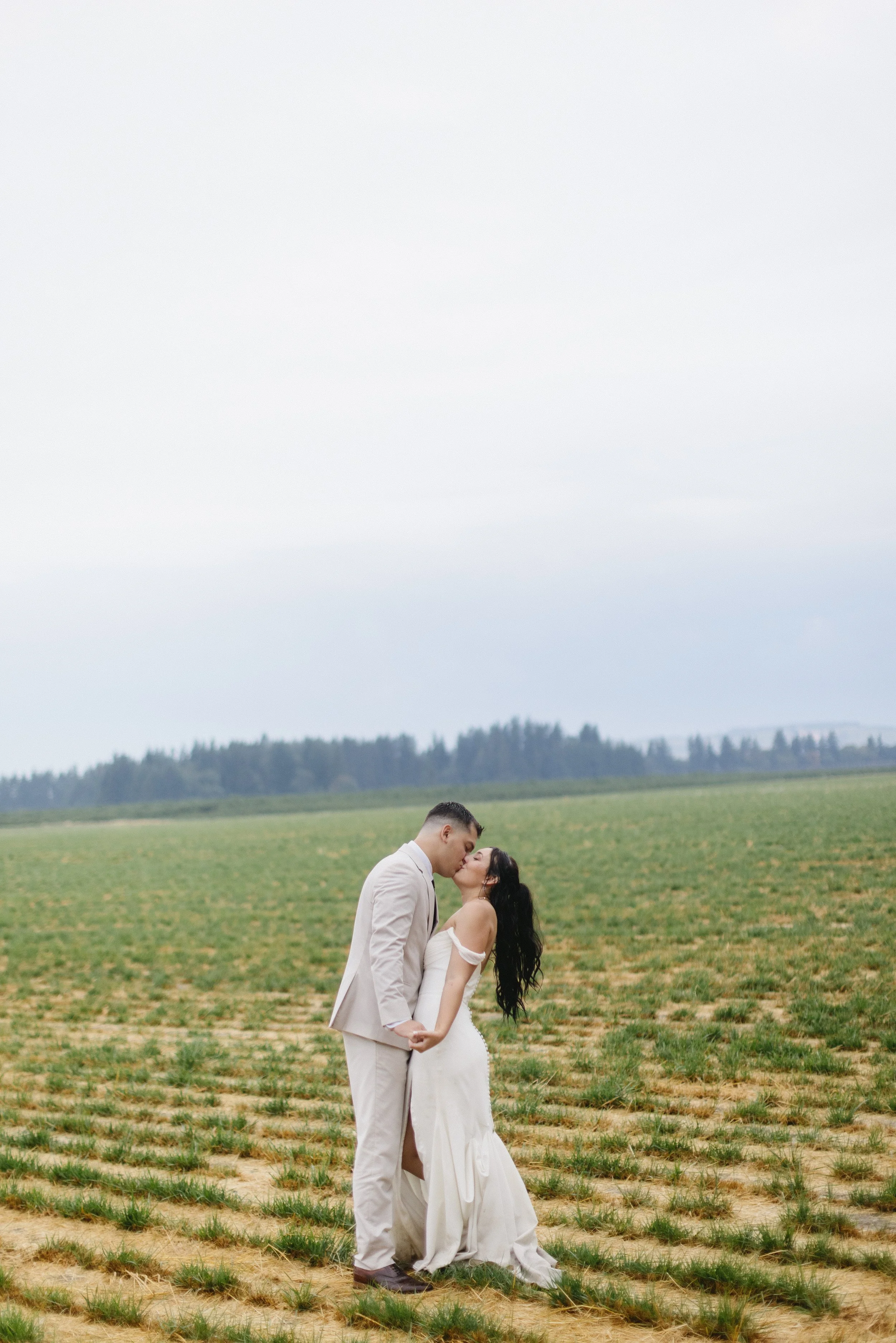 A couple in wedding attire shares a kiss in a grassy field with a distant tree line and overcast sky.