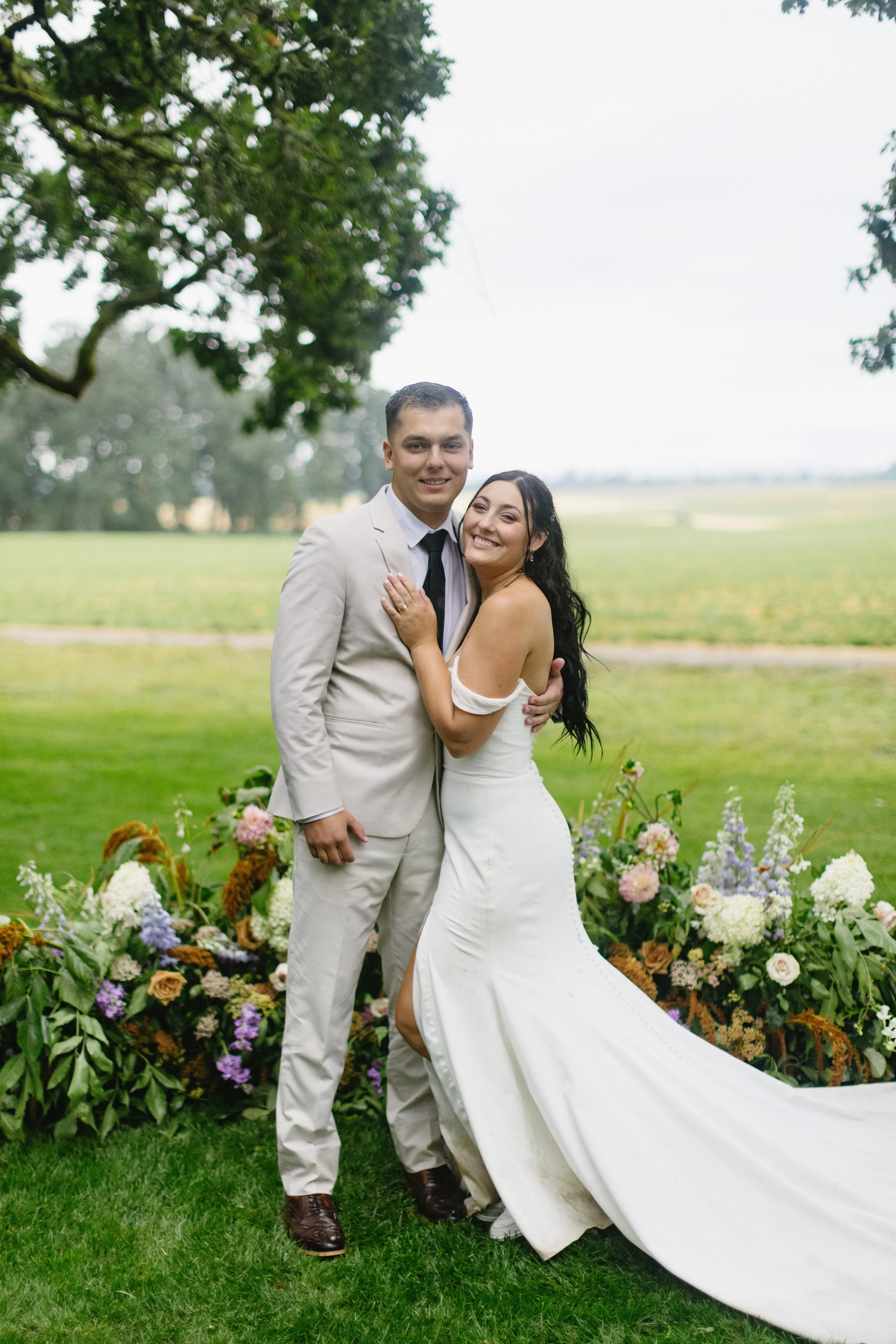 A happy bride and groom standing outdoors on their wedding day, smiling and embracing each other, with a backdrop of flowers and a green field.