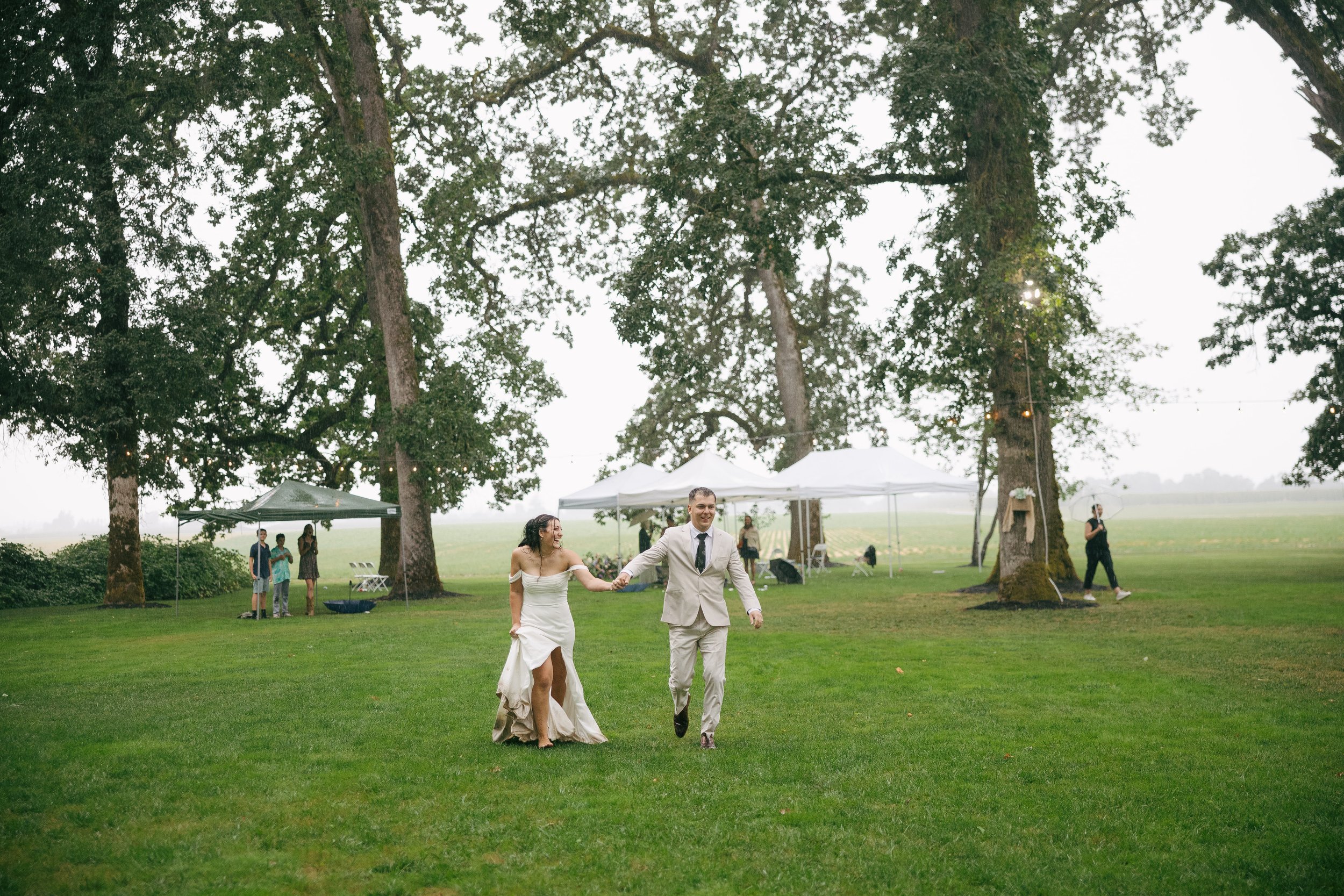 A newlywed couple, a woman in a white wedding dress and a man in a light-colored suit, holding hands and walking on a grassy field during a wedding celebration under large trees with tents nearby. Other people are visible in the background, some unde