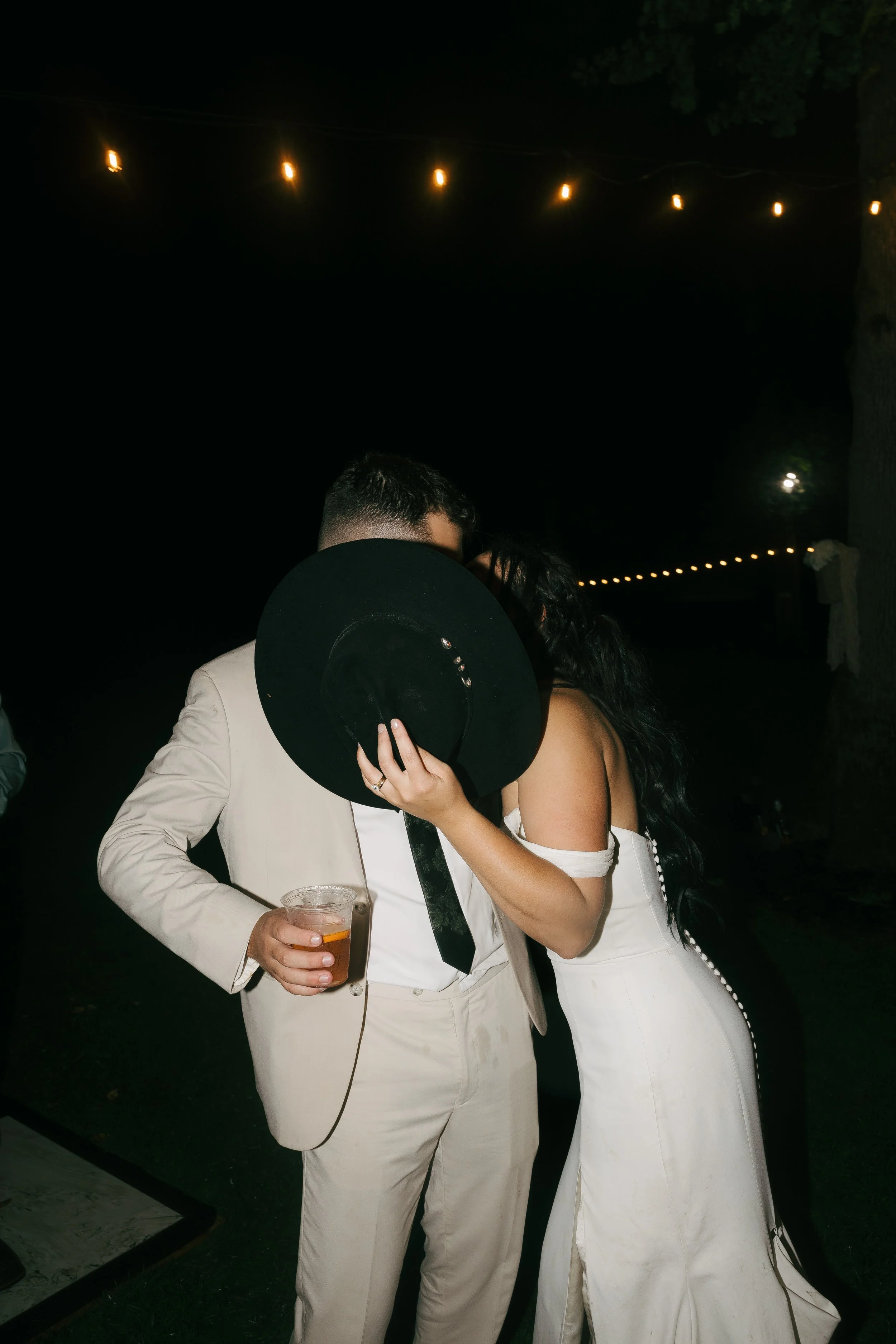 A couple at an outdoor nighttime event, embracing and sharing a kiss, with string lights overhead and one person holding a drink.