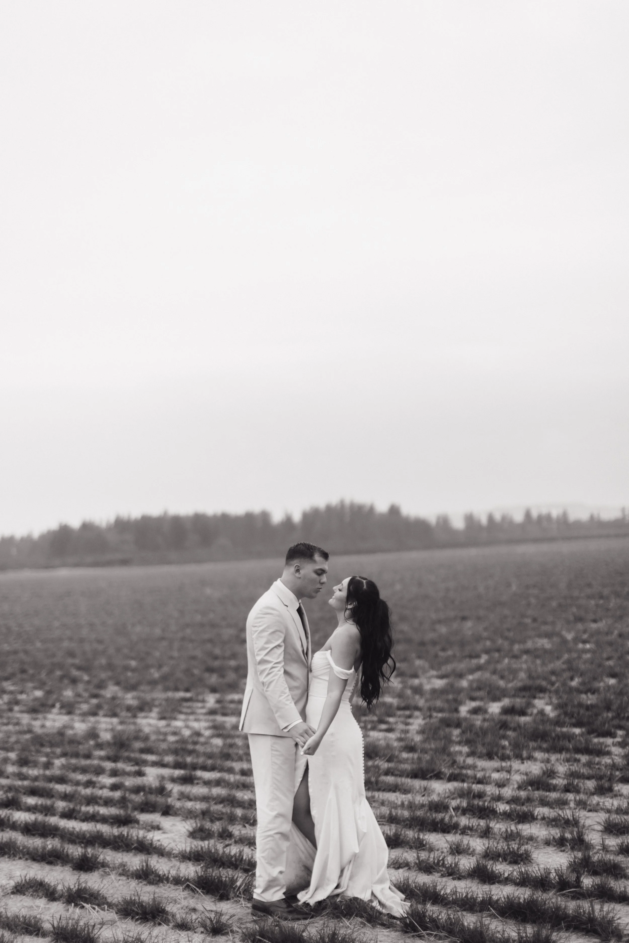 A black-and-white photo of a couple in wedding attire holding hands in an open field, looking at each other.