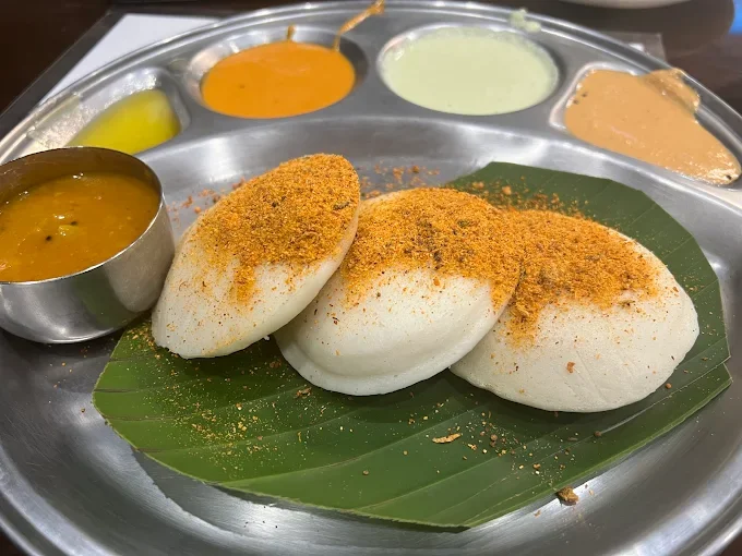 Three steamed idlis sprinkled with ground spices on a banana leaf, served on a metal tray with various chutneys and sambar.