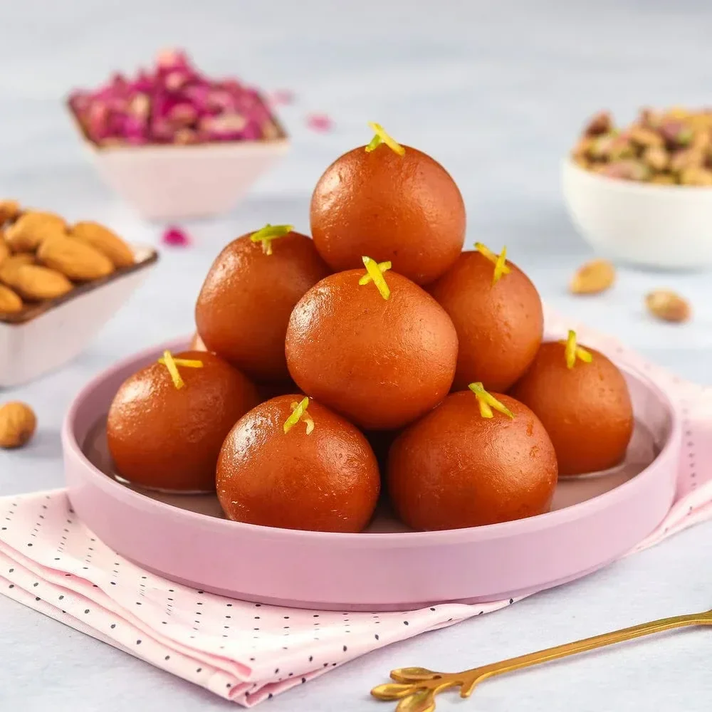A pink plate with a pyramid of Indian sweet treats called gulab jamun, garnished with small yellow edible decorations, with bowls of nuts and dried rose petals in the background.