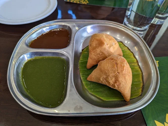 Two samosas on a banana leaf inside a stainless steel plate, with green and brown sauces on the side.