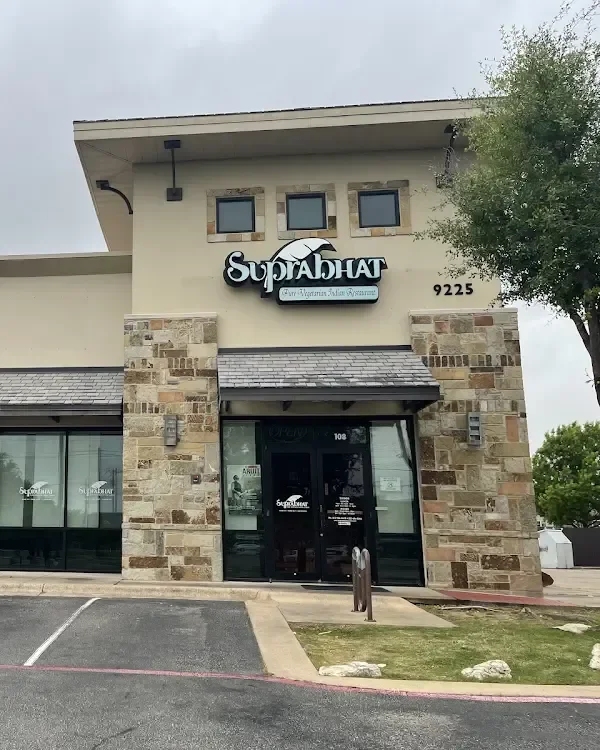Outside of a sushi restaurant called SupaHut with a sign showing a fish and umbrella, located at 9225, with a stone and stucco facade, parking lot, trees, and cloudy sky.