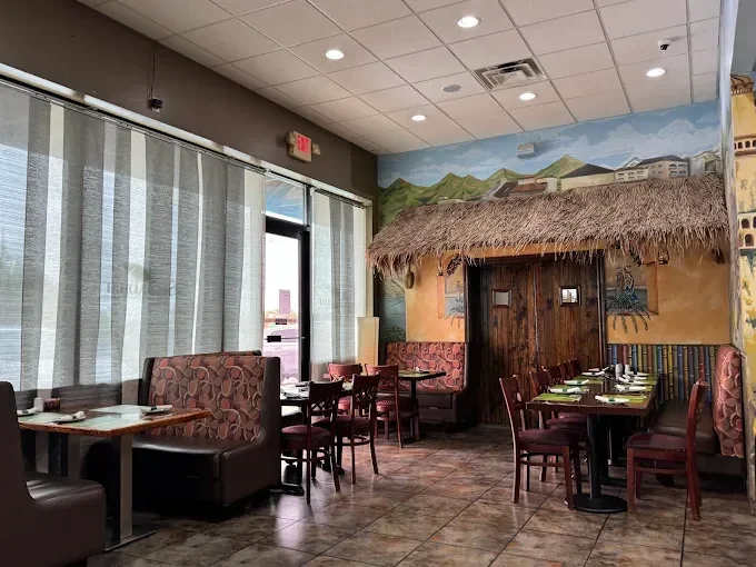 Interior of a restaurant with painted mountain scenery, a thatched hut feature, and mixed seating including booths and chairs, with natural light coming through large window with curtains.