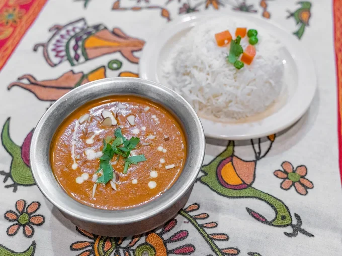 Plate of white rice garnished with vegetables and a bowl of Indian curry topped with herbs on a decorative tablecloth.