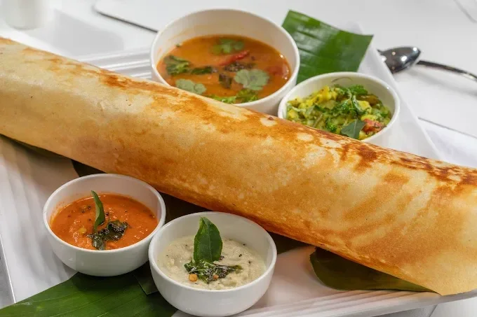 Indian meal featuring a large crispy dosa with five small bowls of different chutneys and sambar on a white tray.