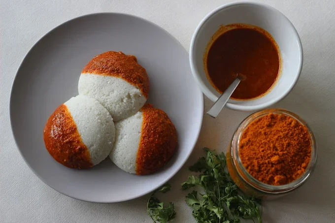 Three white and brown Indian-style steamed rice cakes called idlis on a white plate, a bowl of spicy red chutney, a jar of bright orange spice powder, and some fresh green cilantro on a white table