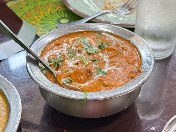 A bowl of Indian curry garnished with cilantro, with a spoon inside, on a dark wooden table, alongside a glass of water and a colorful plate.