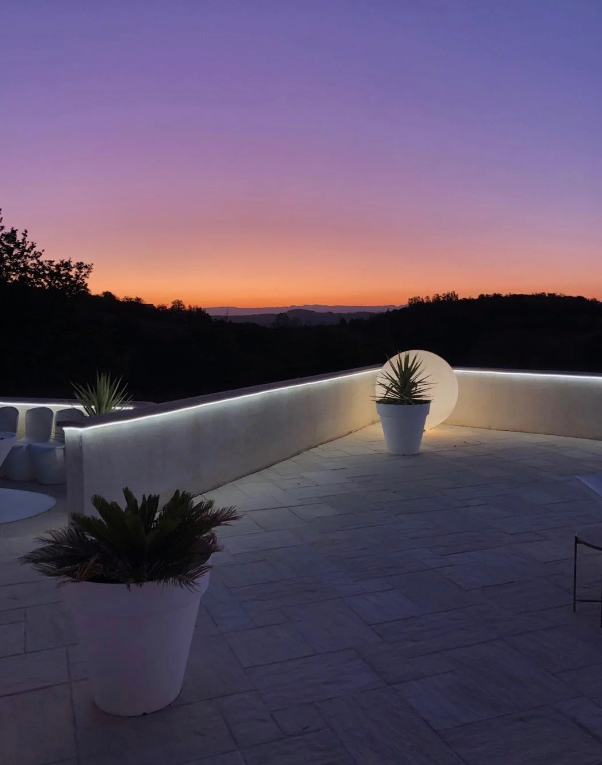 Conception d'éclairages pour une terrasse moderne avec des pots de plantes et des lumières LED, vue sur un paysage au coucher du soleil avec un ciel violet et orange.