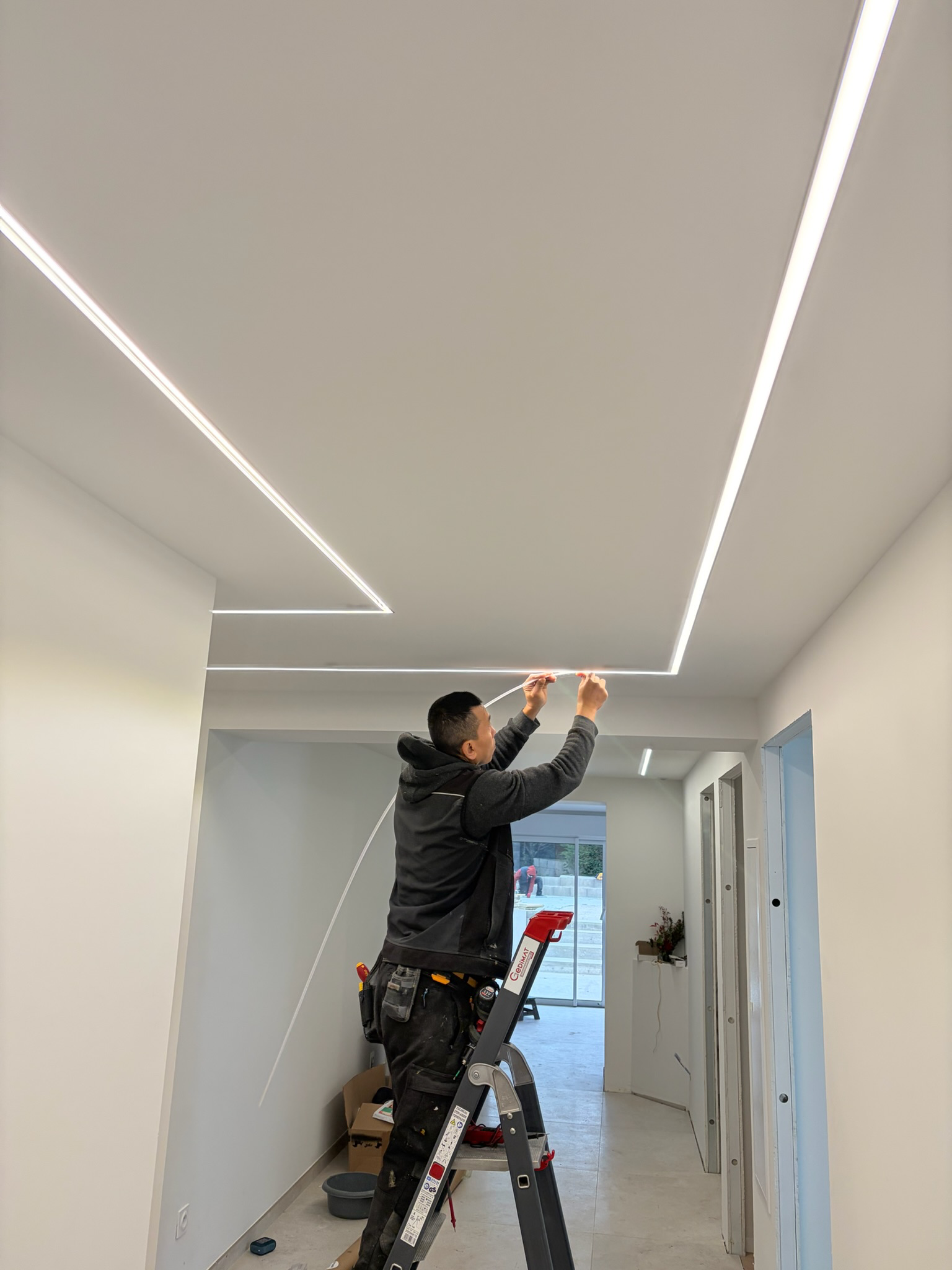 Un homme monte une échelle pour installer un éclairage LED encadré dans un plafond blanc, dans un intérieur moderne.