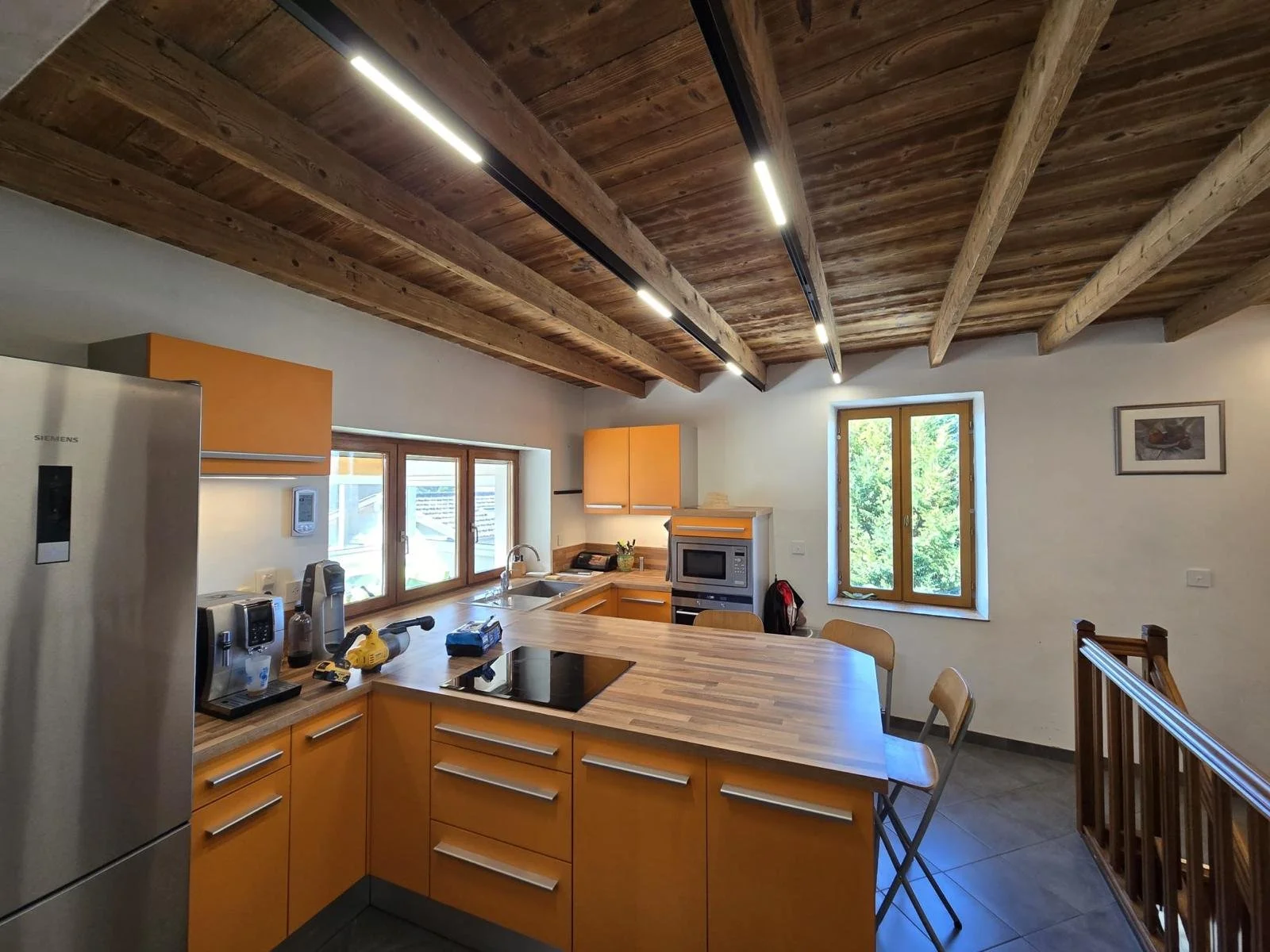 Conception d'éclairages pour une cuisine moderne avec comptoir en bois, armoires orange, appareils électroménagers en acier inoxydable, fenêtres avec vue sur la nature, plafond en bois avec poutres apparentes.