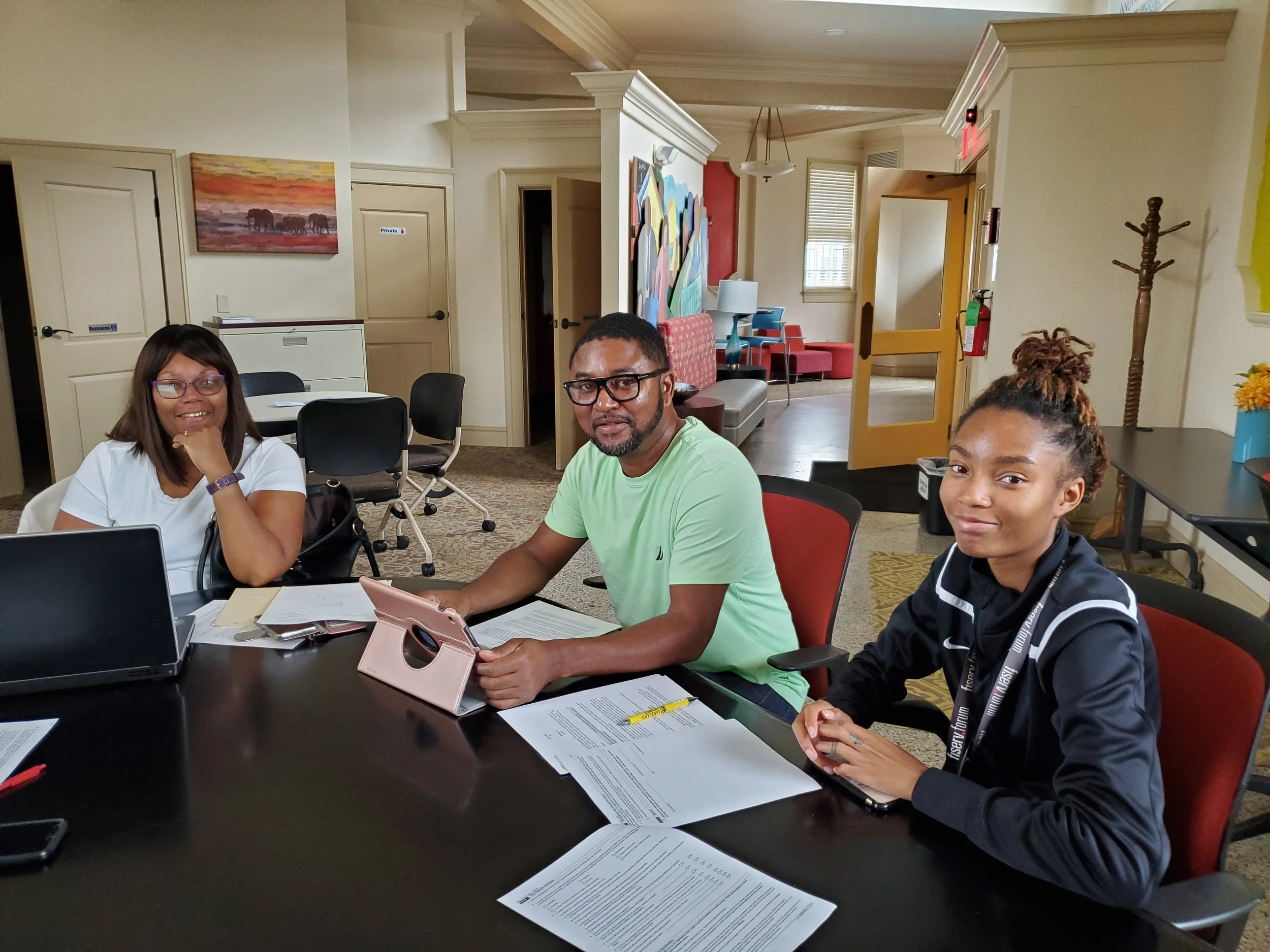 Three people sitting at a conference table in a meeting room, with documents, laptops, and a tablet. The woman on the left has glasses and a white shirt, the man in the center has glasses and a green shirt, and the young woman on the right has a curly updo hairstyle and a black jacket.