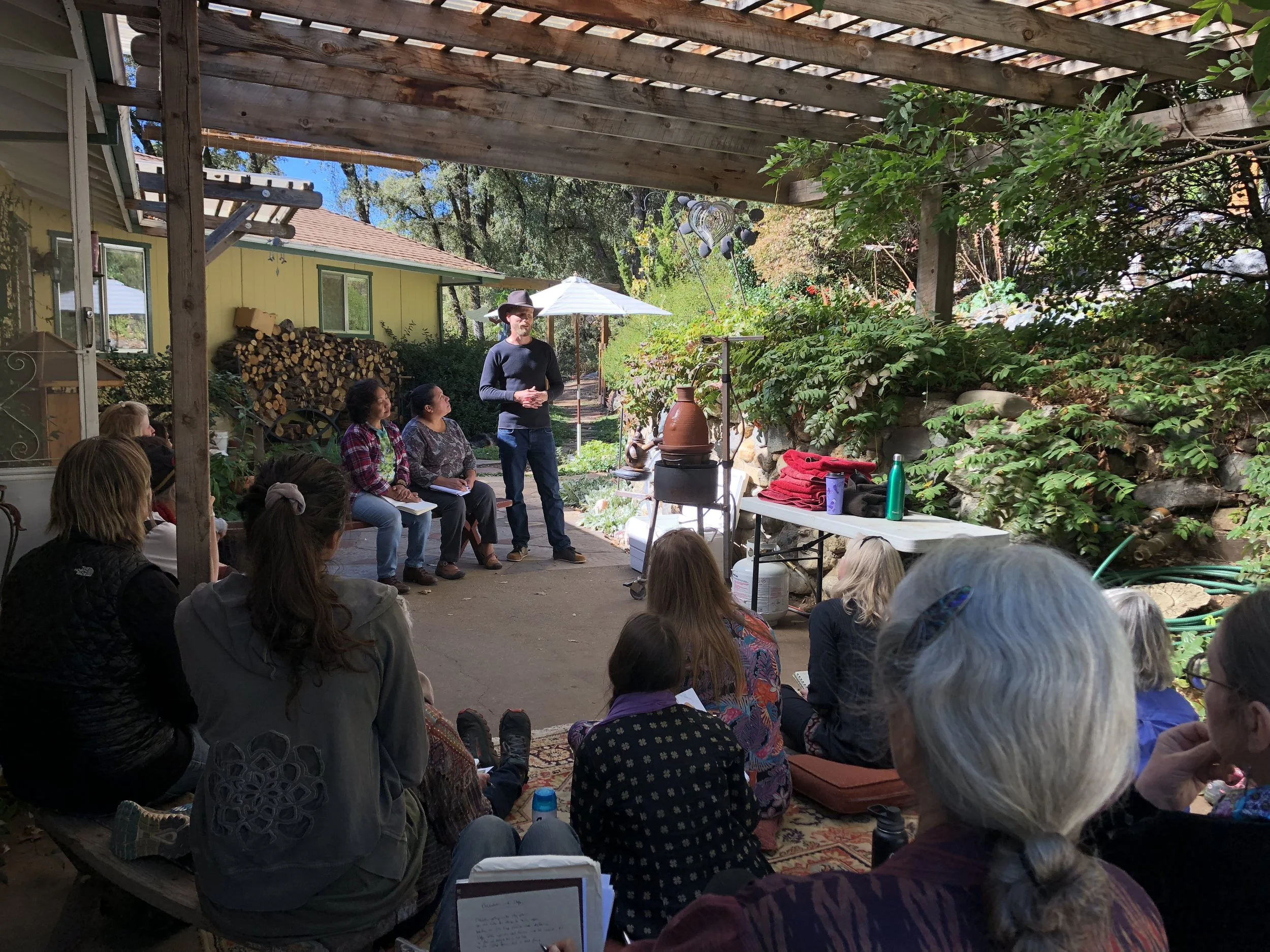 A group of people gathered outdoors under a wooden roof, listening to a man in a black shirt and hat giving a presentation. The setting is a garden with greenery, a yellow house, and stacked firewood in the background.