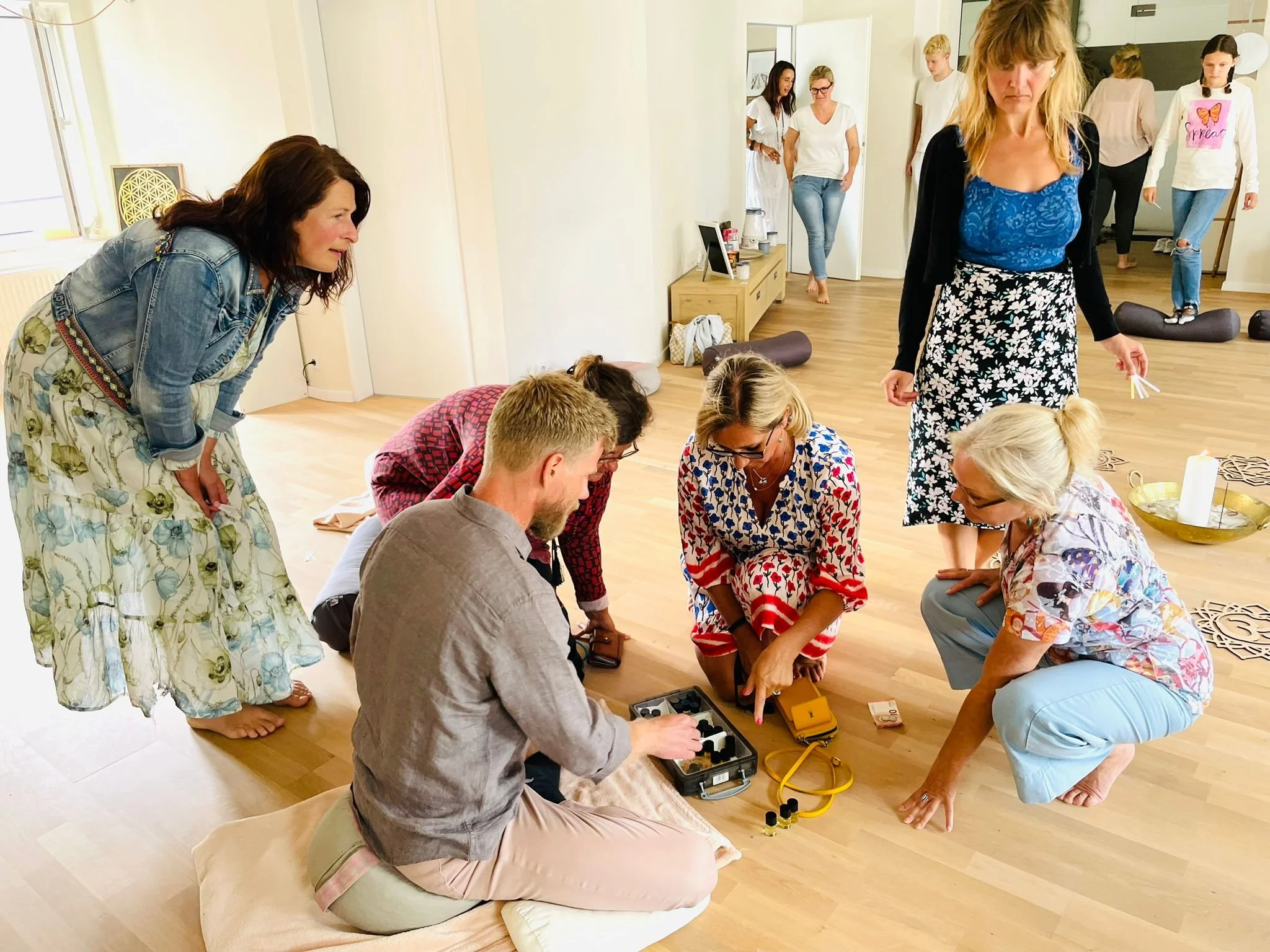 Group of women and one man practicing aromatherapy or essential oils session on a wooden floor in a bright room, some participants kneeling or sitting, others standing, with scent bottles and diffuser.