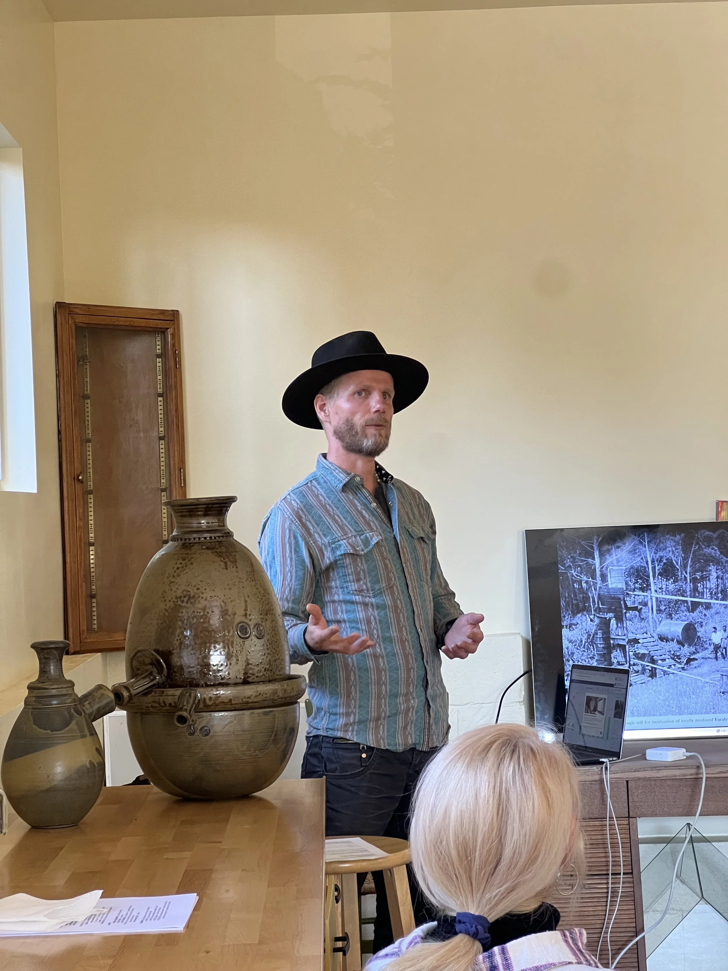 A man wearing a black hat and a striped shirt is giving a presentation in a room. There are pottery vases on a table nearby and a woman with blonde hair is seated and listening. A large screen displays an old black-and-white photograph of a rural scene.
