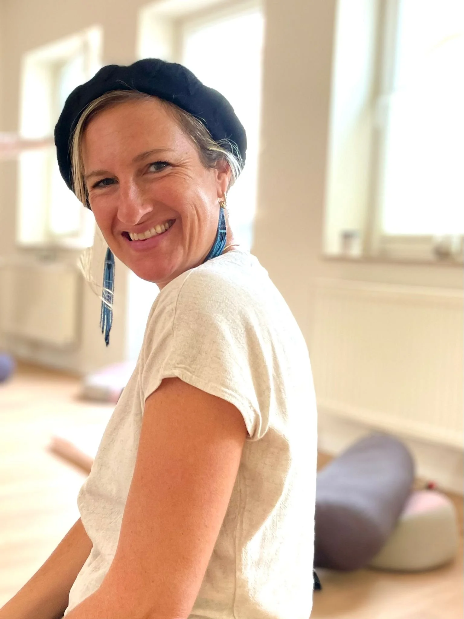 Smiling woman with blonde hair, wearing a black beret and a white shirt inside a well-lit room with windows.