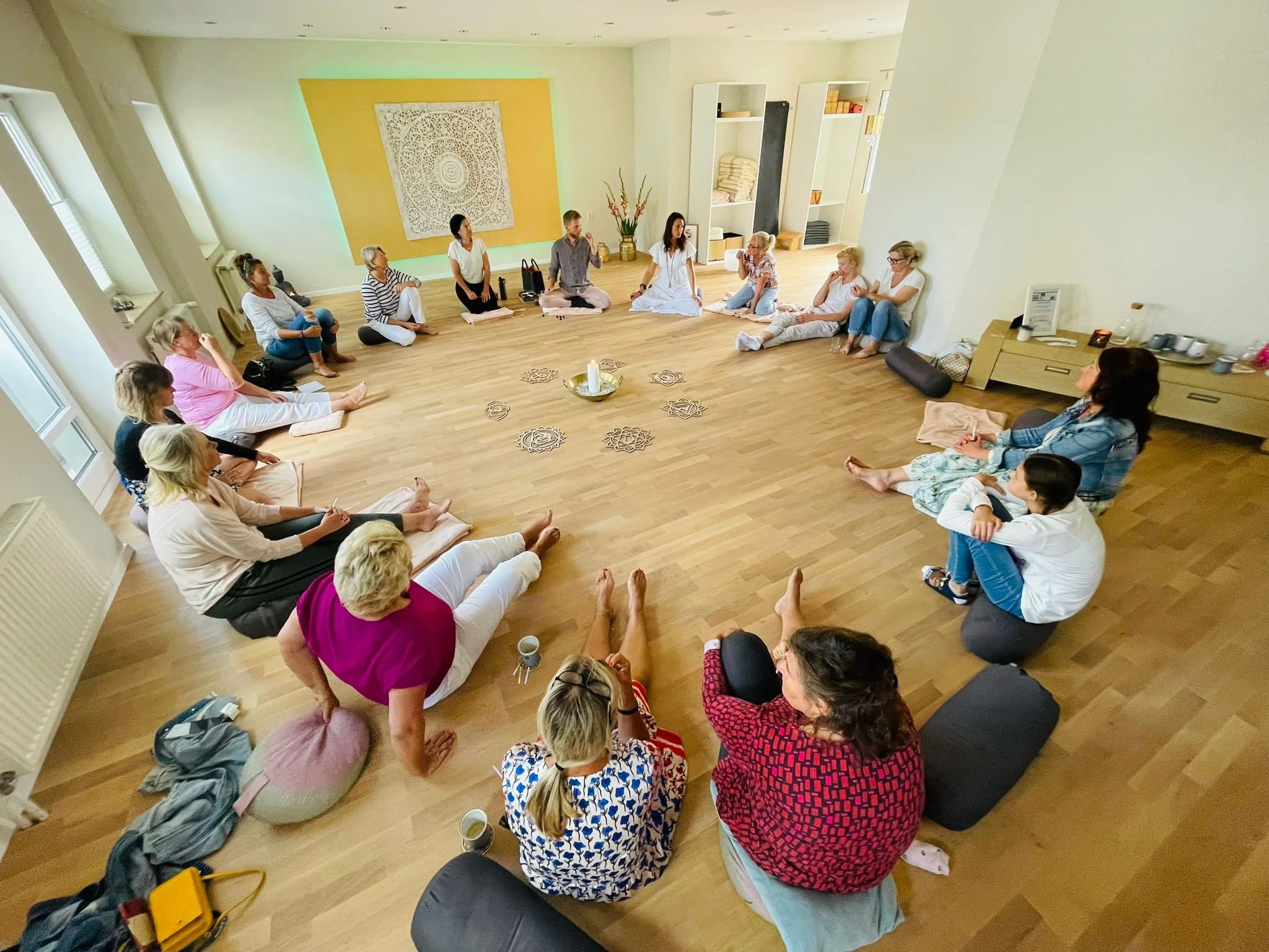 Group of people sitting in a circle on the floor of a spacious room, participating in a group activity or meditation session, with a small table holding a candle and decorative items in the center.
