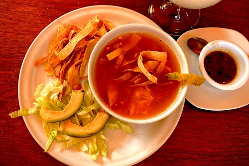 Bowl of clear sopa de lima with tortilla strips beside avocado and a small dish of salsa