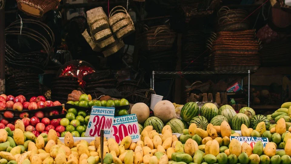 Fruit and baskets on a Mexico City market stall, with handwritten peso price cards among mangoes and melons under warm indoor light.