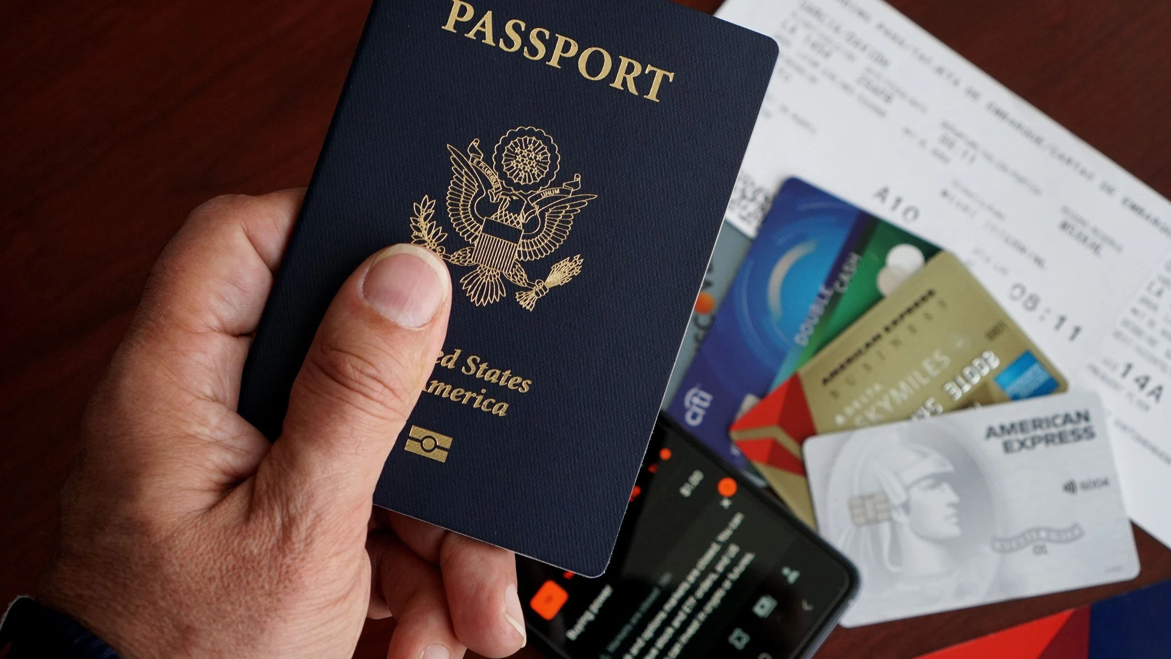 A hand holds a United States passport over bank cards, a phone and a printed ticket on a table