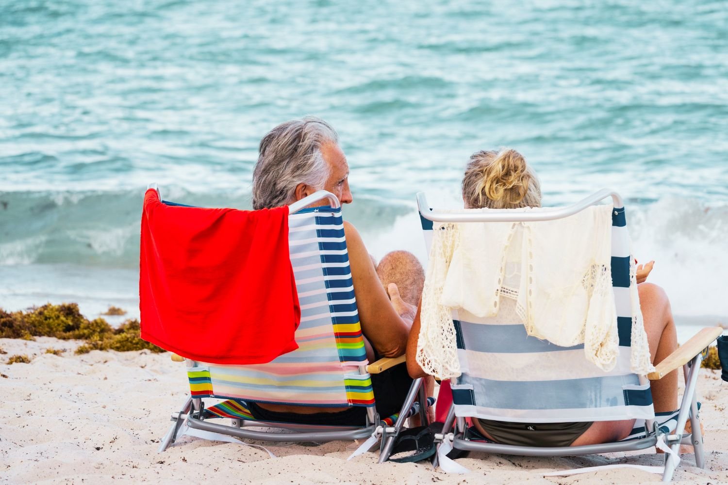 Retired couple sitting on beach loungers by the sea on white sand.
