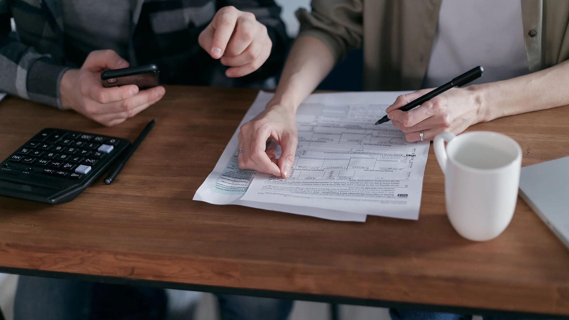 Two people's hands going through a blank US tax form at a wooden table with a calculator and mug