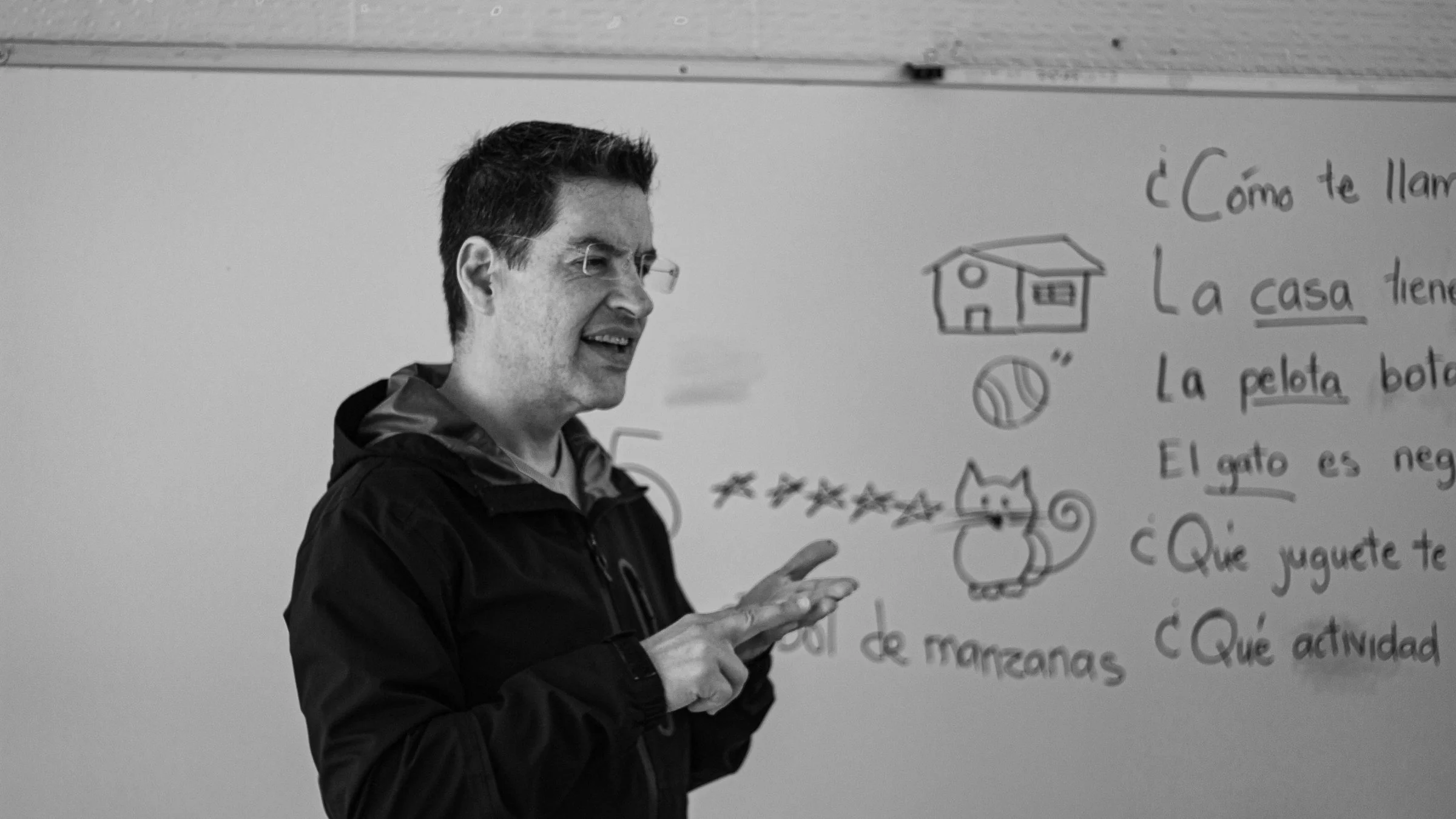 A Spanish teacher gestures beside a whiteboard of simple Spanish sentences and sketches, shown in black and white