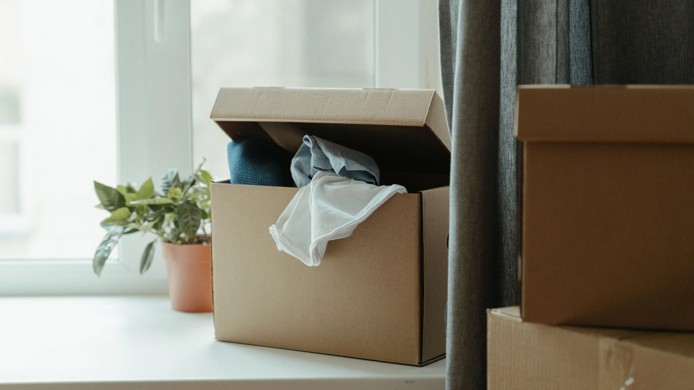 An open cardboard box of clothes, a second box and a potted plant on a bright windowsill beside a grey curtain