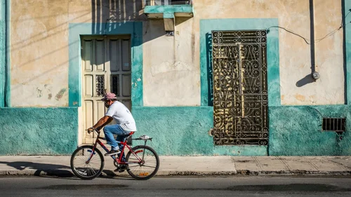 Cyclist riding past a weathered teal and cream colonial house with ironwork doors in Mérida