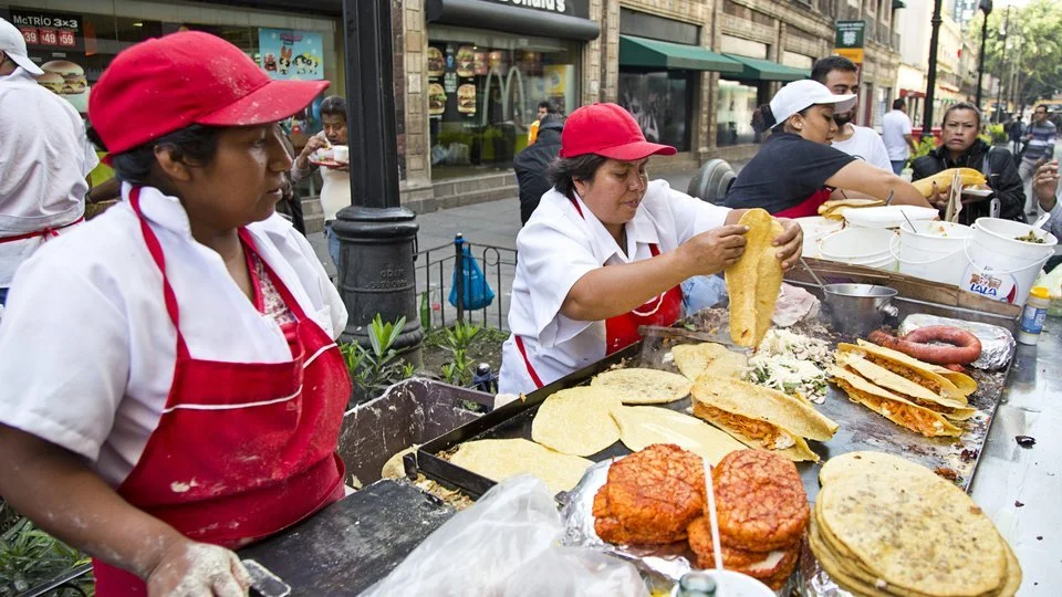 Street food stall in Mexico City with cooks in red aprons preparing quesadillas and tortillas on a large griddle on the pavement, with city shopfronts behind.