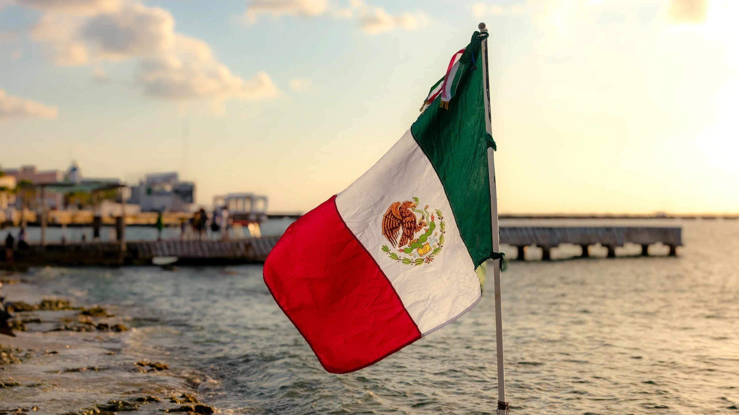 A Mexican flag flutters by the sea in Cozumel, Mexico, with a pier and waterfront buildings in soft sunset light