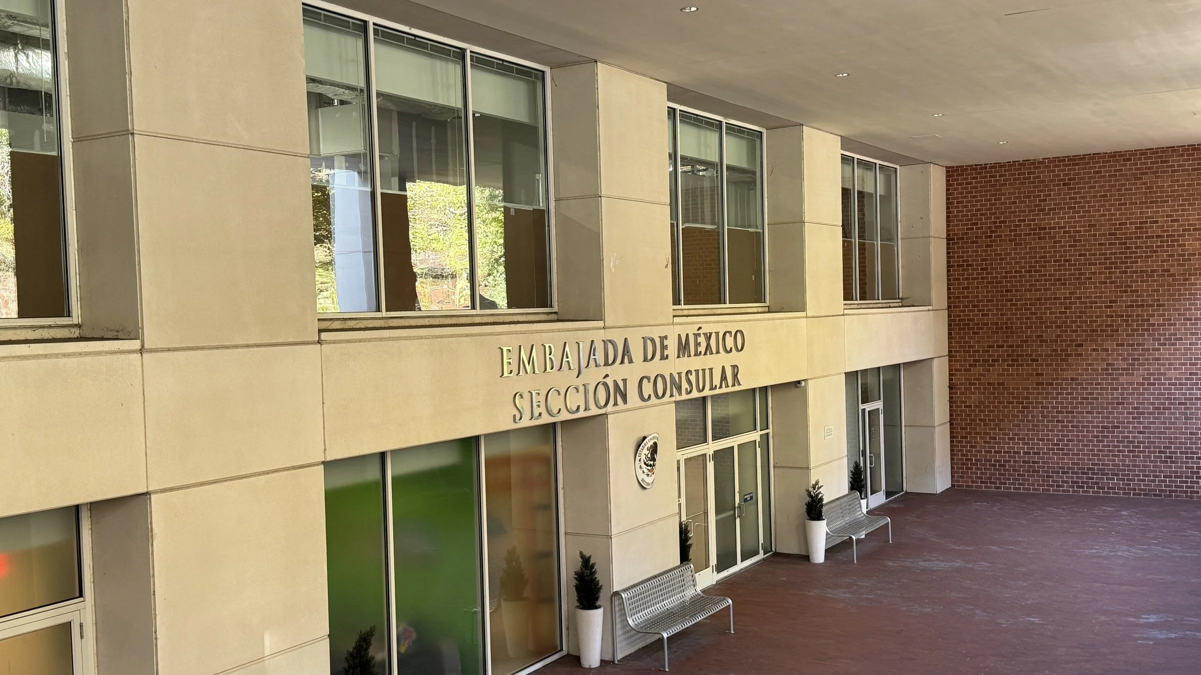 Entrance to the consular section of the Embassy of Mexico, with metal lettering reading Embajada de México, Sección Consular, a circular Mexican coat of arms, glass doors and metal benches under a covered walkway.