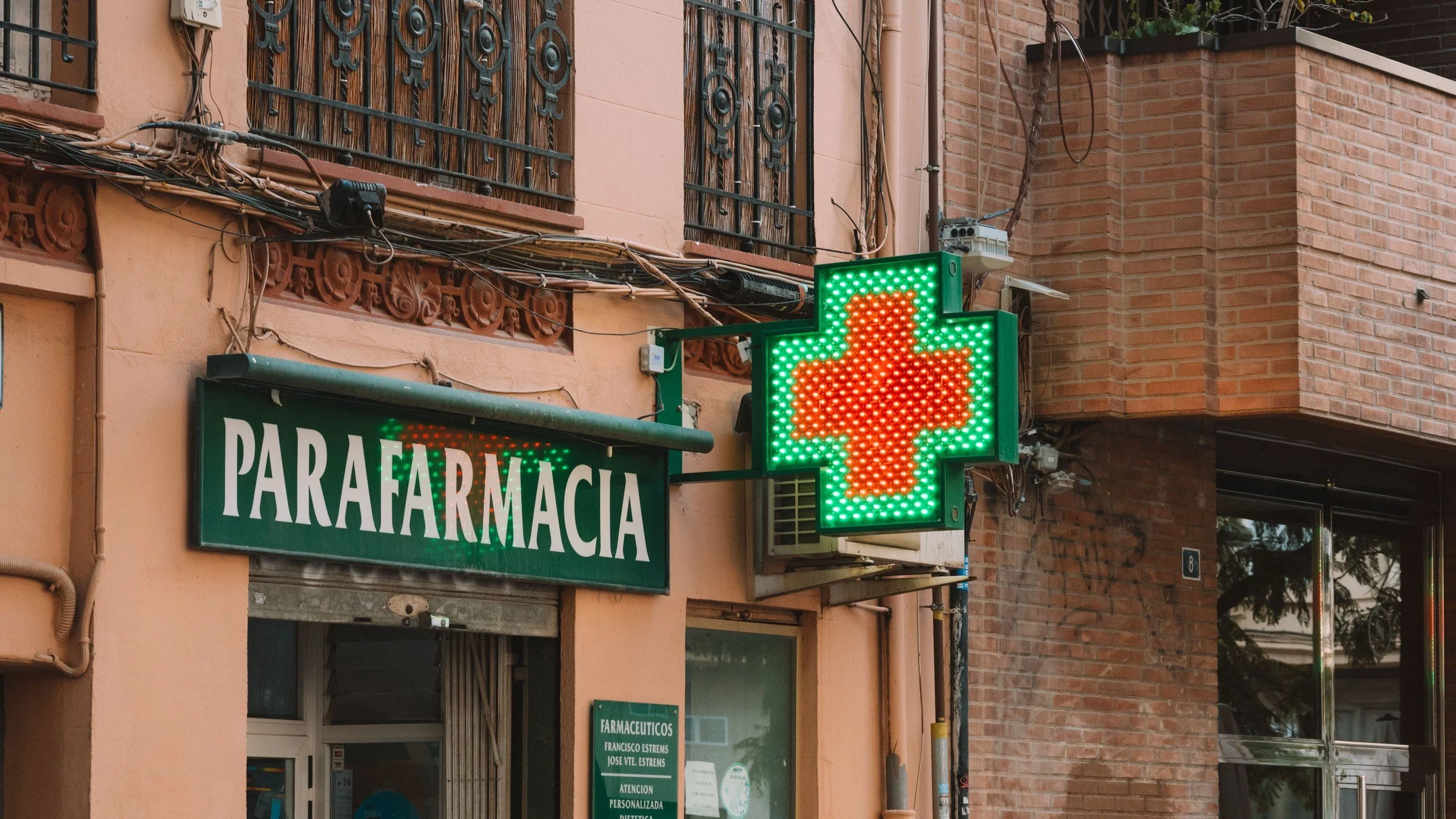 Pharmacy shopfront with a glowing green cross sign above the door on a sunlit street