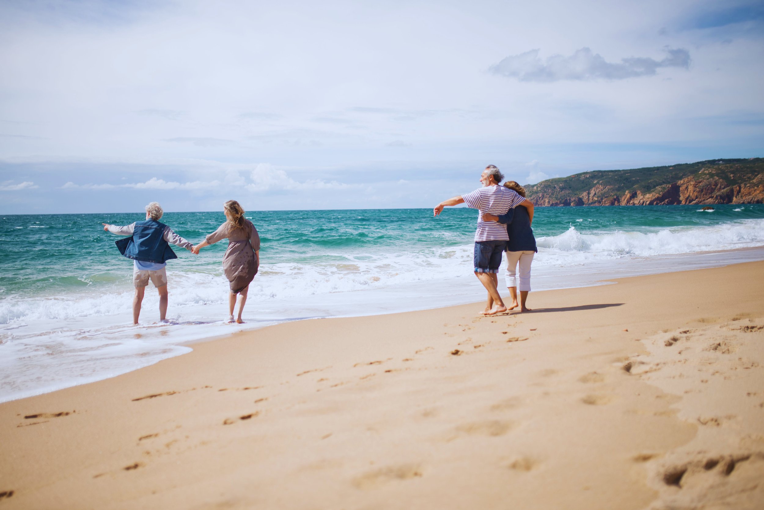 Four people walk on a sandy beach near the ocean, holding hands and enjoying the sunny weather with clouds in the sky and a coastal hill in the background.