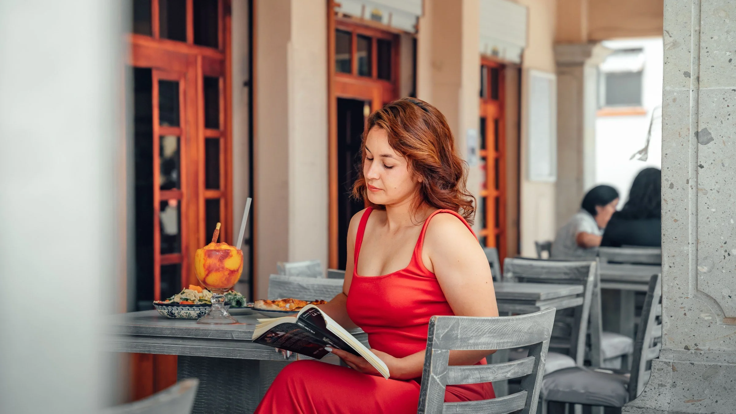 A woman in a red dress reads a book at a café table under a stone arcade in Lerma, State of Mexico