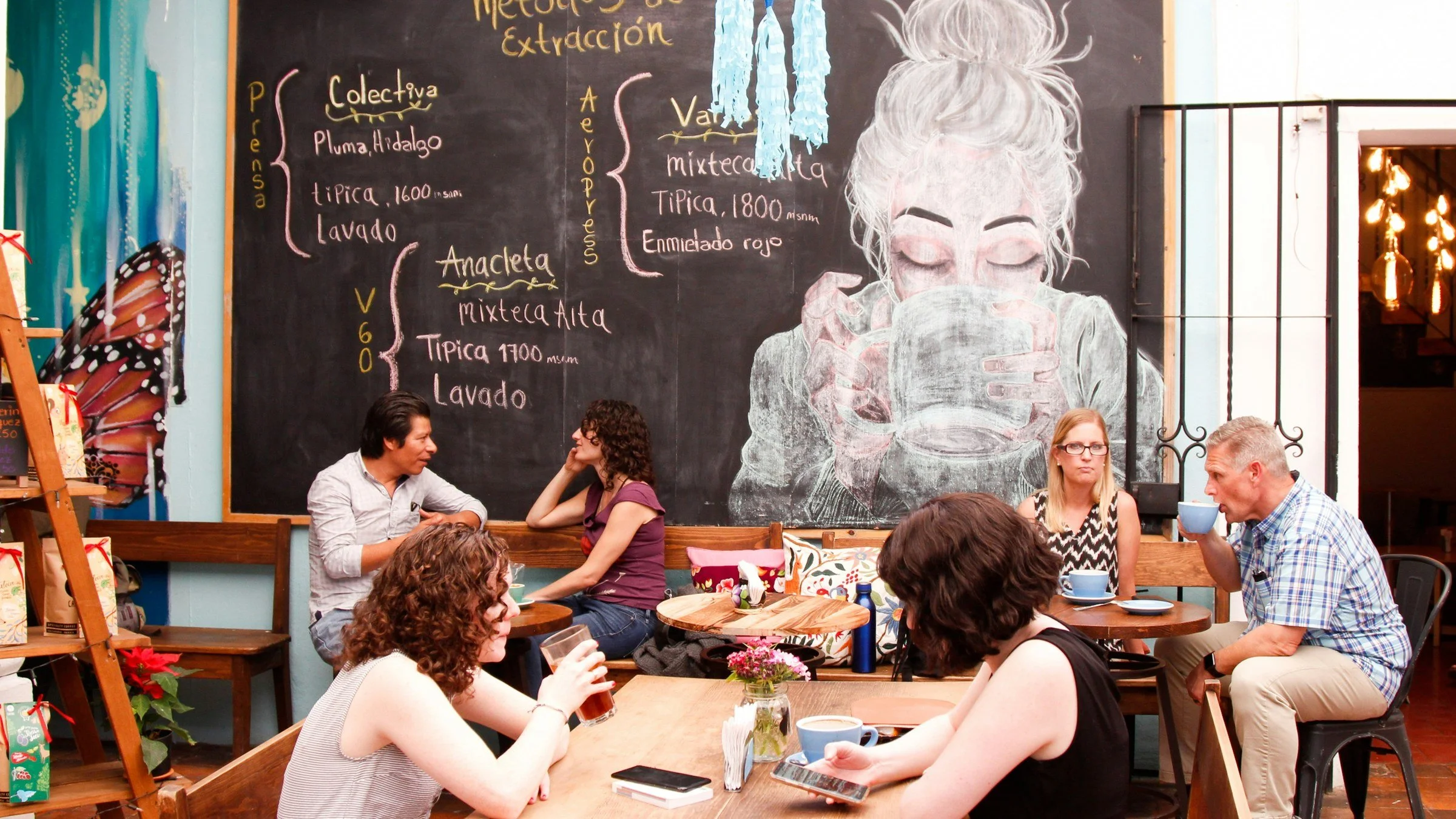 Adults chat over coffee at wooden tables in a café in Oaxaca, beneath a chalkboard coffee menu written in Spanish