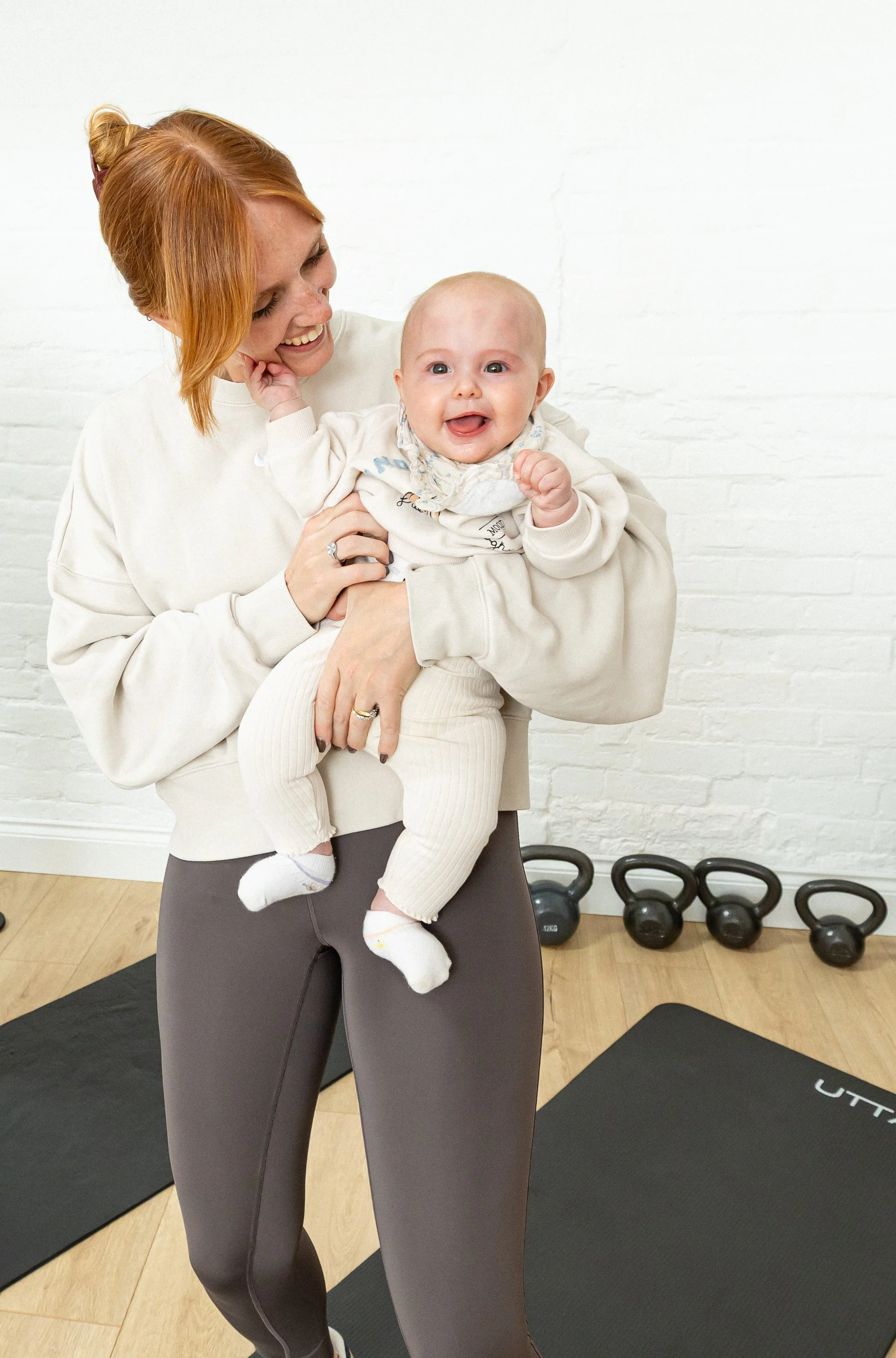 A woman holding a baby in a fitness class studio with kettlebells and exercise mats in the background.