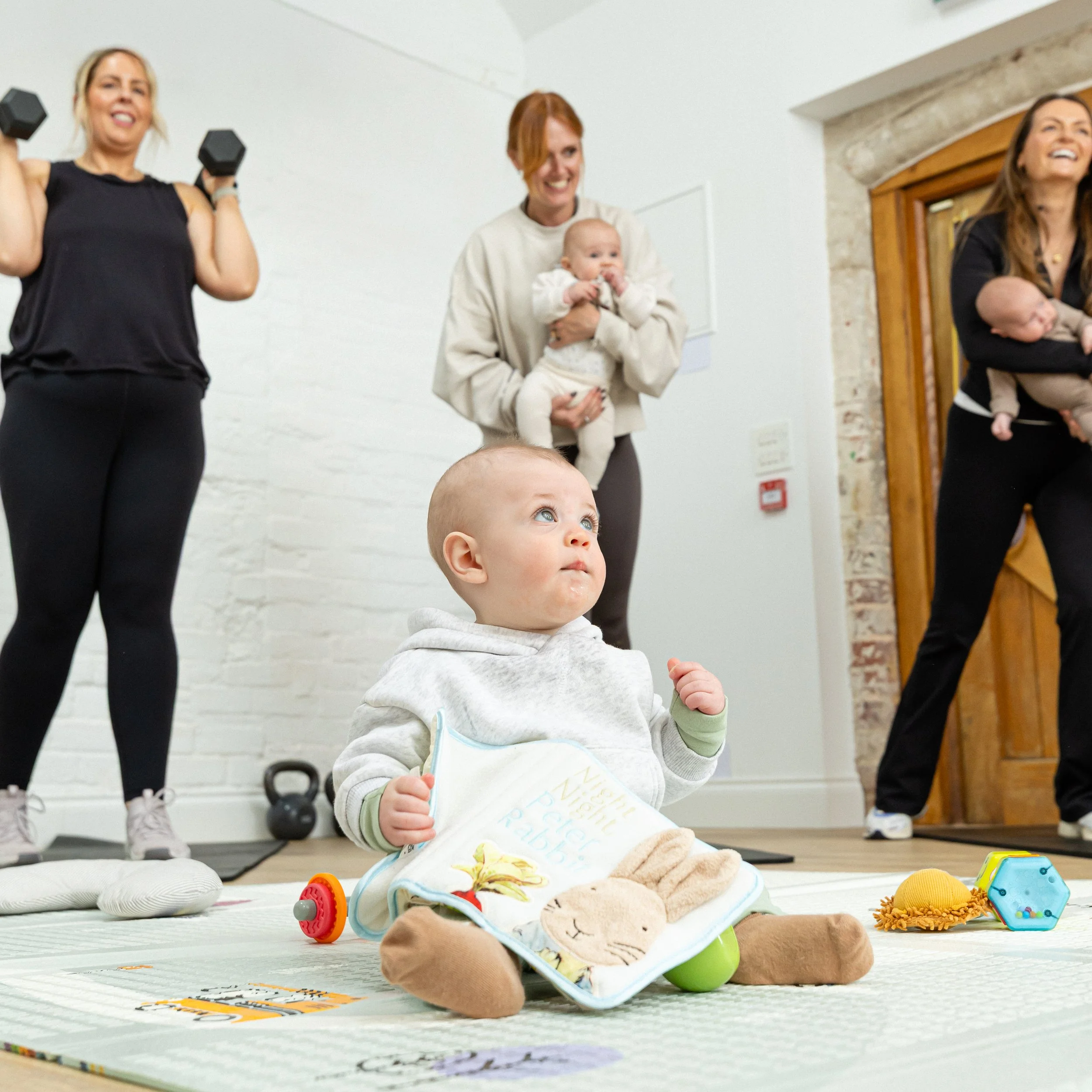 A young child sitting on a play mat with toys scattered around, wearing a gray hoodie with green cuffs and khaki pants, looking up. In the background, three women are smiling, two holding babies and one lifting dumbbells.