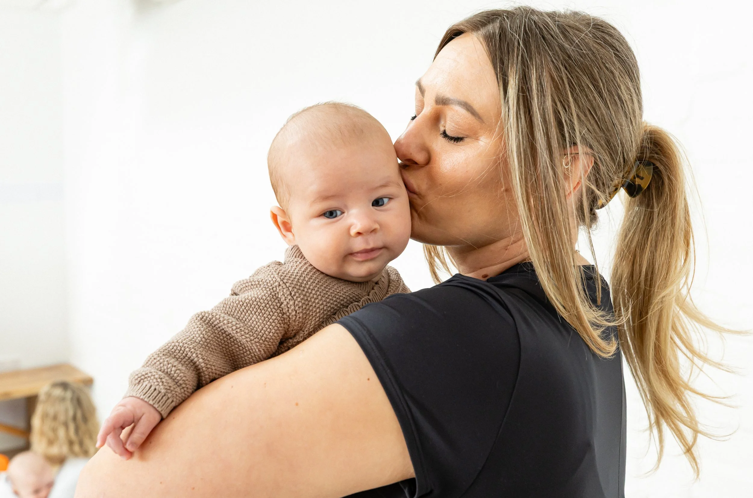 A woman with long blonde hair in a ponytail is holding and kissing a baby on the forehead. The baby has light skin, blue eyes, and is wearing a brown, textured sweater, looking slightly to the side. The background is a plain, light-colored wall.