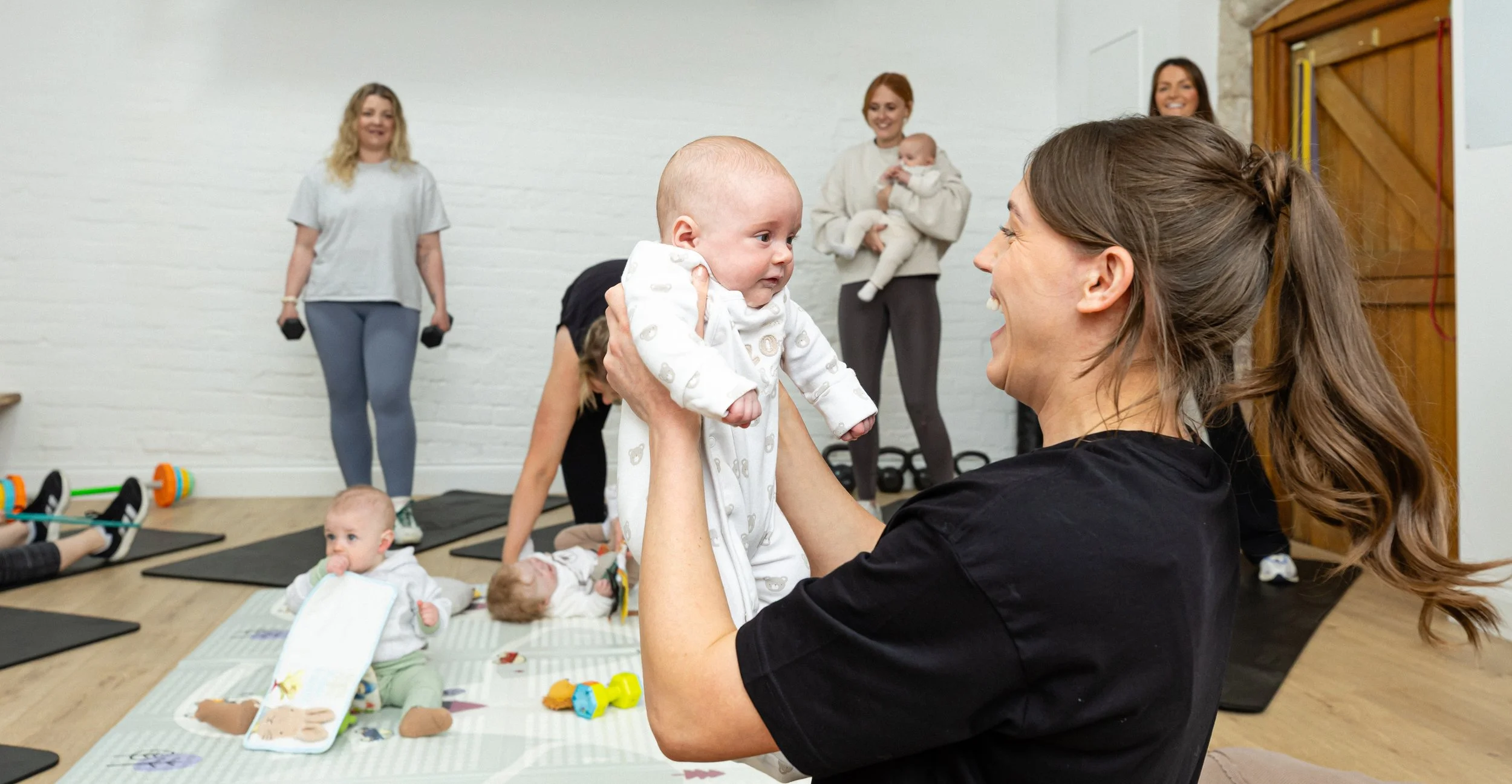 A woman is smiling and holding a baby while doing a workout class. Several other children are lying on mats or sitting nearby, and a few women are standing in the background in a gym or studio with white brick walls.