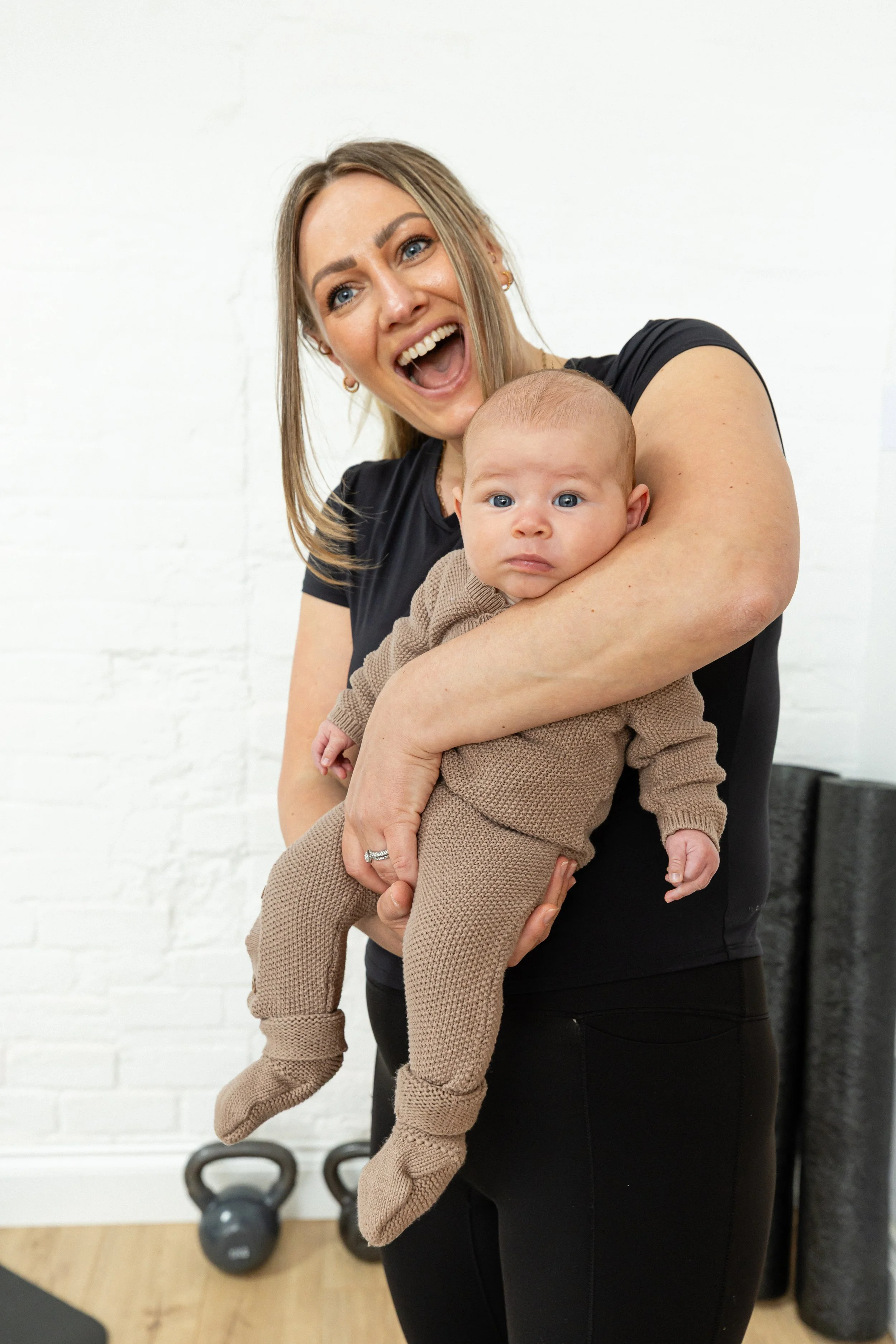 A woman with blonde hair and blue eyes holding a baby with light brown hair and blue eyes, both smiling, in a fitness studio with kettlebells and black foam rollers.