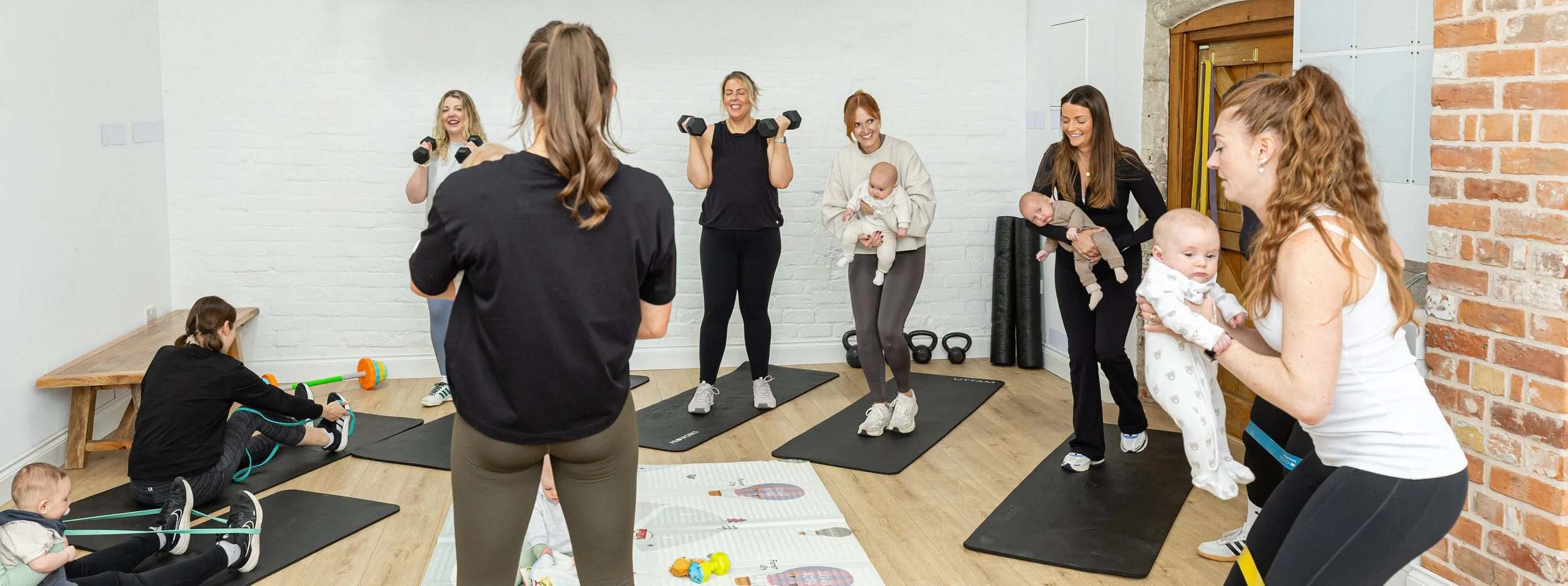Women participating in a fitness class with babies and young children in a room with white brick walls and wood flooring.
