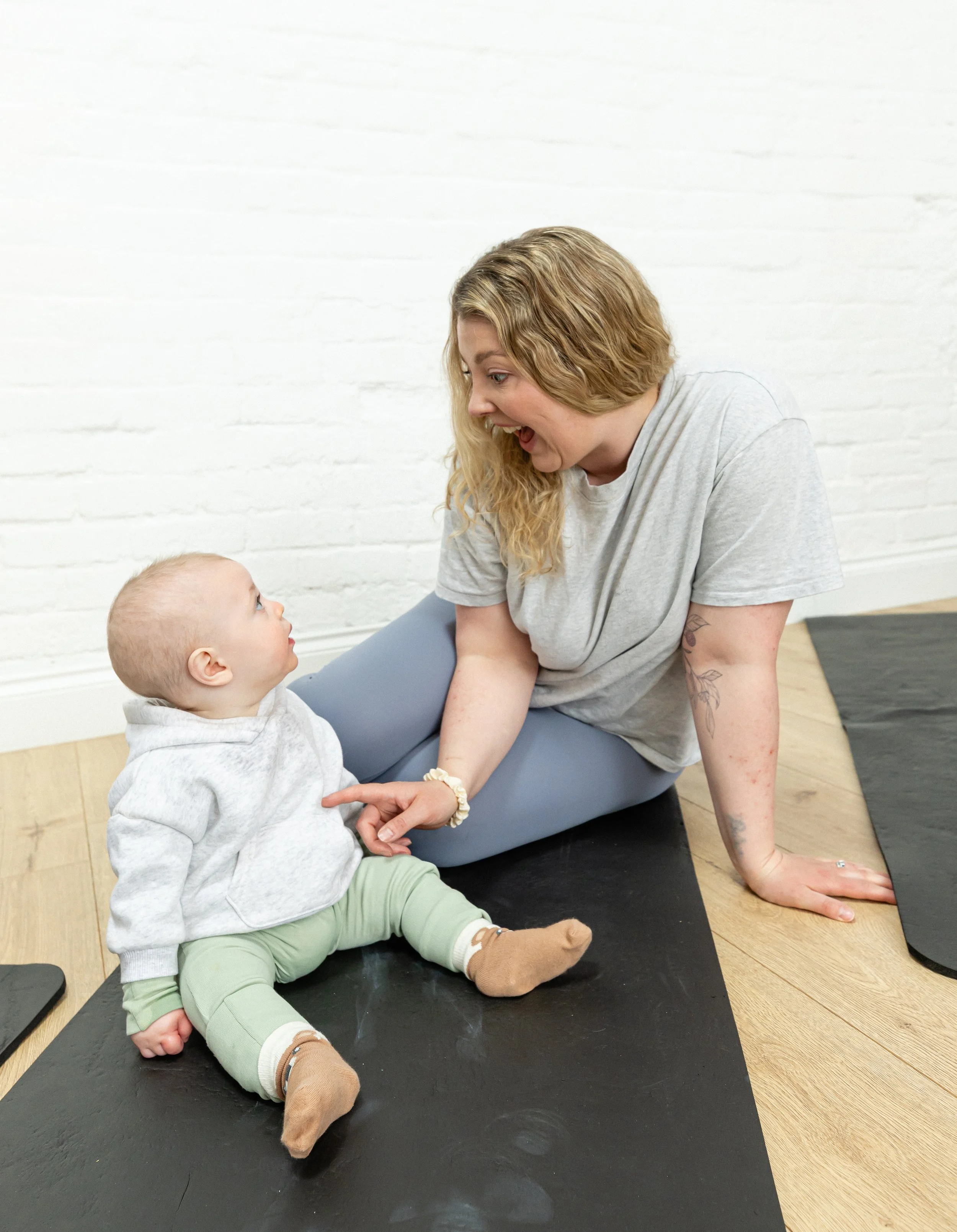 A woman with blonde curly hair, wearing a gray T-shirt, is engaging with a baby sitting on a black yoga mat. The baby has light blond hair, is dressed in a gray hoodie, green pants, and brown socks. They are in a room with white brick walls and wooden flooring, and there is a gray yoga mat in the background.