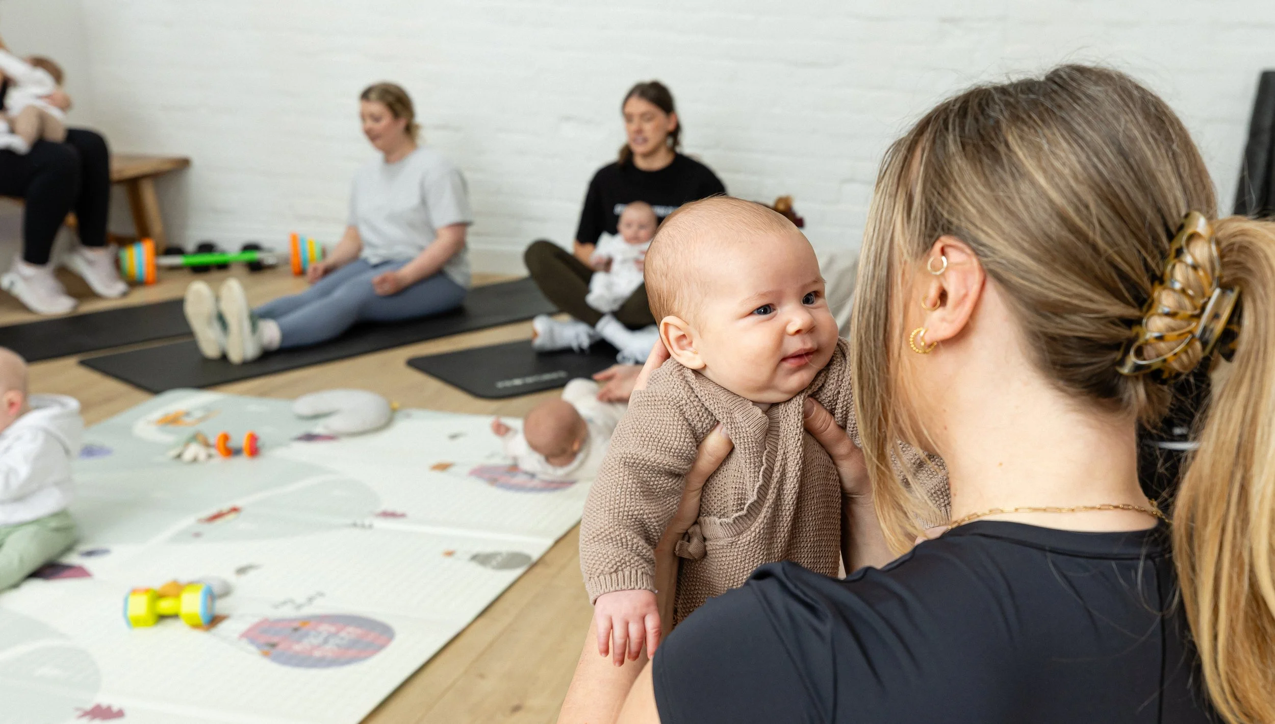 A woman with blonde hair in a ponytail and a black shirt holds a baby close to her face during a baby activity class. In the background, other women engage with babies and toys on mats in a room with white brick walls.