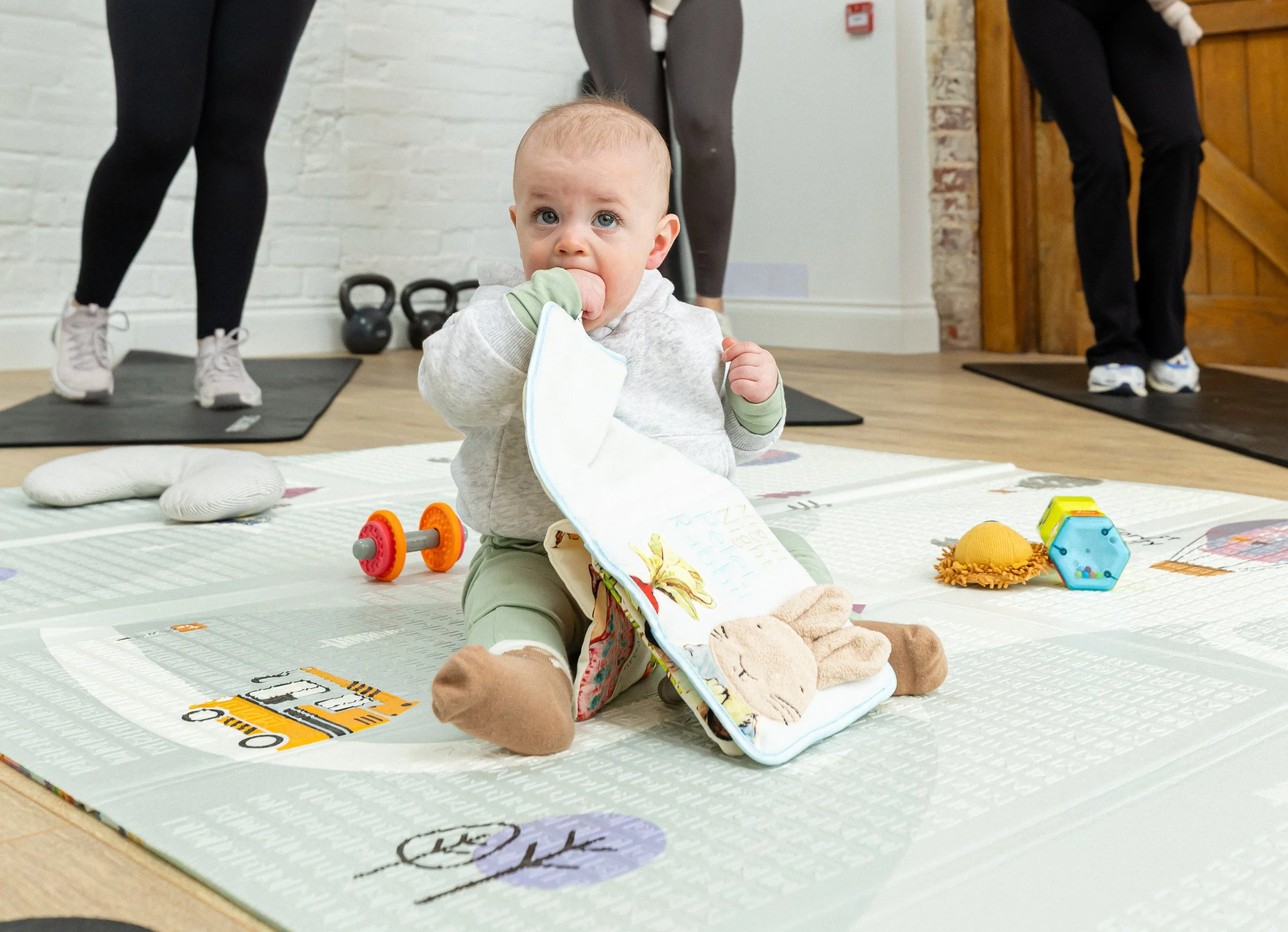 A baby sitting on the floor in a gym or exercise studio, sucking on their hand, with adults in workout clothes standing behind, and various toys and toys around the baby.